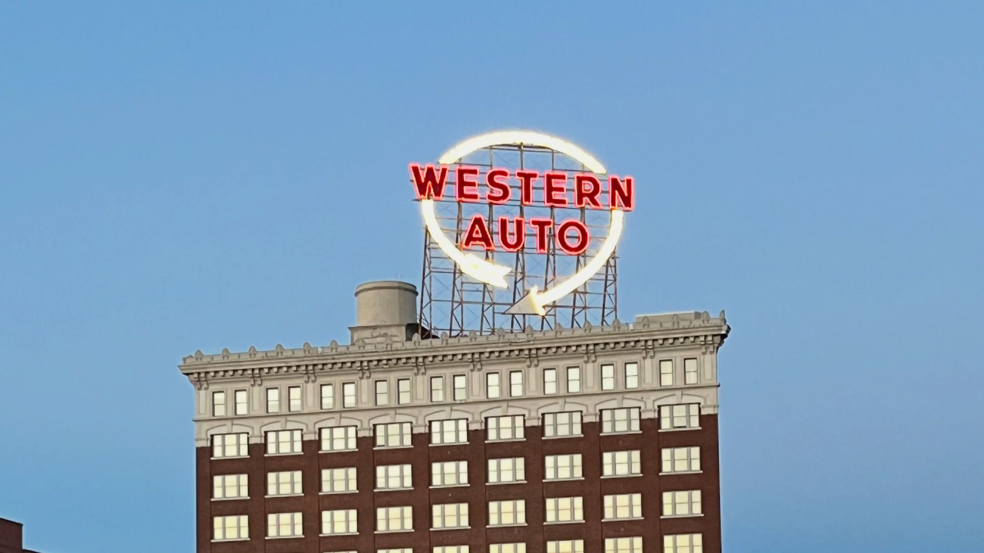 Tall brick building with a large neon red WESTERN AUTO sign encircled by a glowing ring atop, set against a clear blue sky.