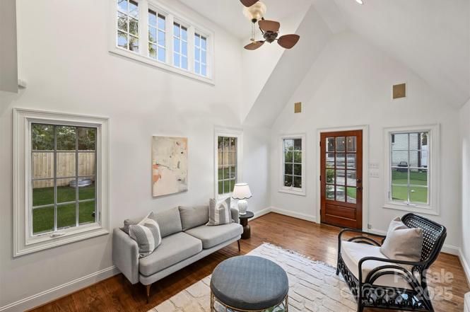 Bright living room with vaulted ceiling, white walls, wood floor, gray sofa, round gray ottoman, black rattan chair with cushion, wood door, and multiple windows showing backyard.