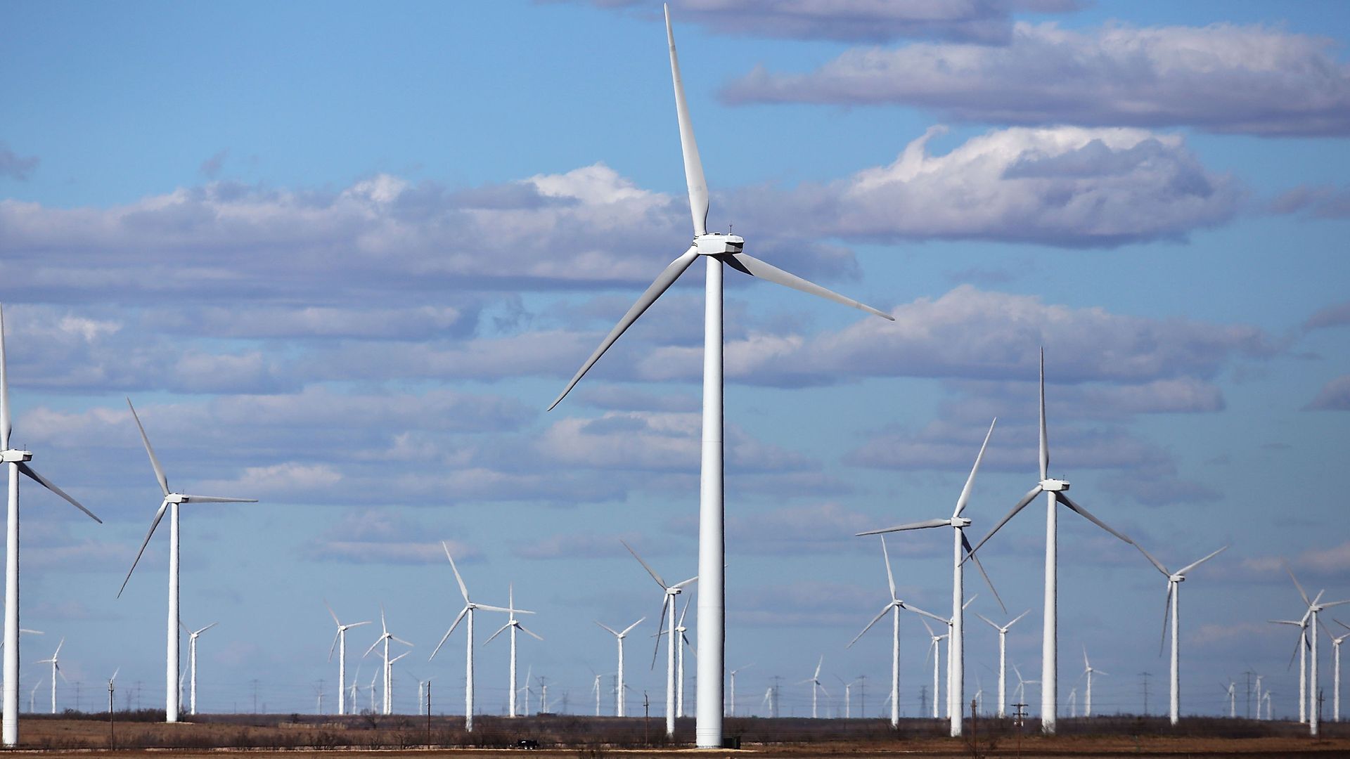 A truck passes by wind turbines in Texas, set against a blue sky with clouds