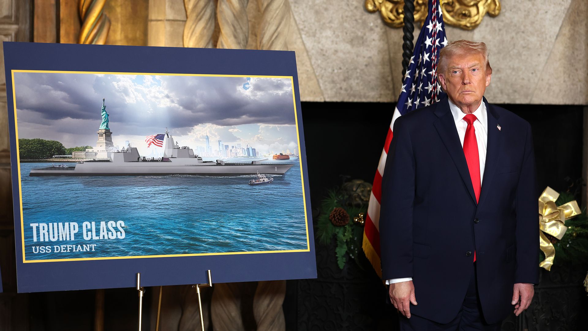 President Trump in a dark suit and red tie, standing beside a large blue poster showing a naval ship and the Statue of Liberty with an American flag.