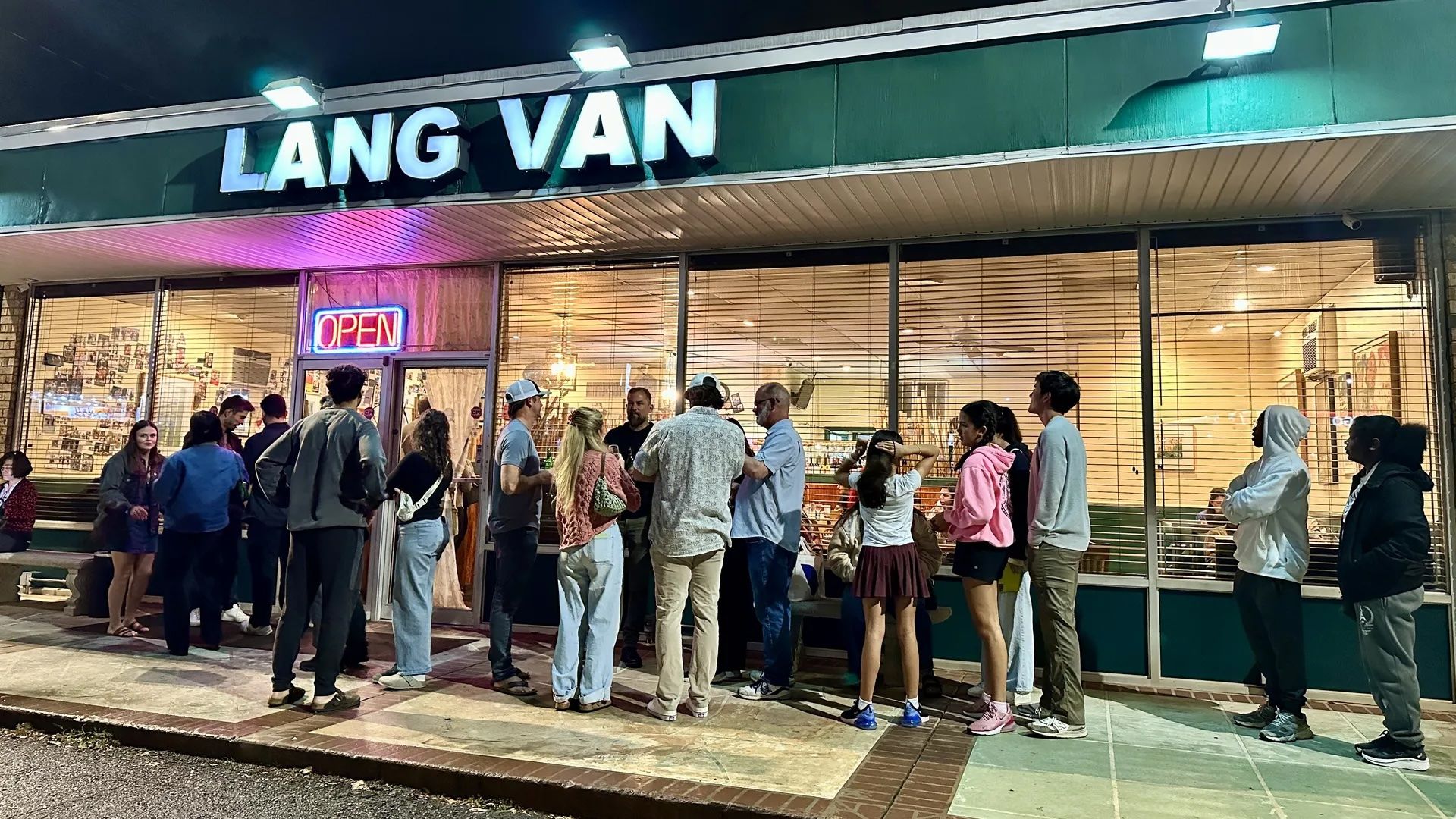 Night view of a crowd waiting outside Lang Van store with an illuminated "OPEN" sign, large front windows with blinds, and green exterior walls under bright lights.
