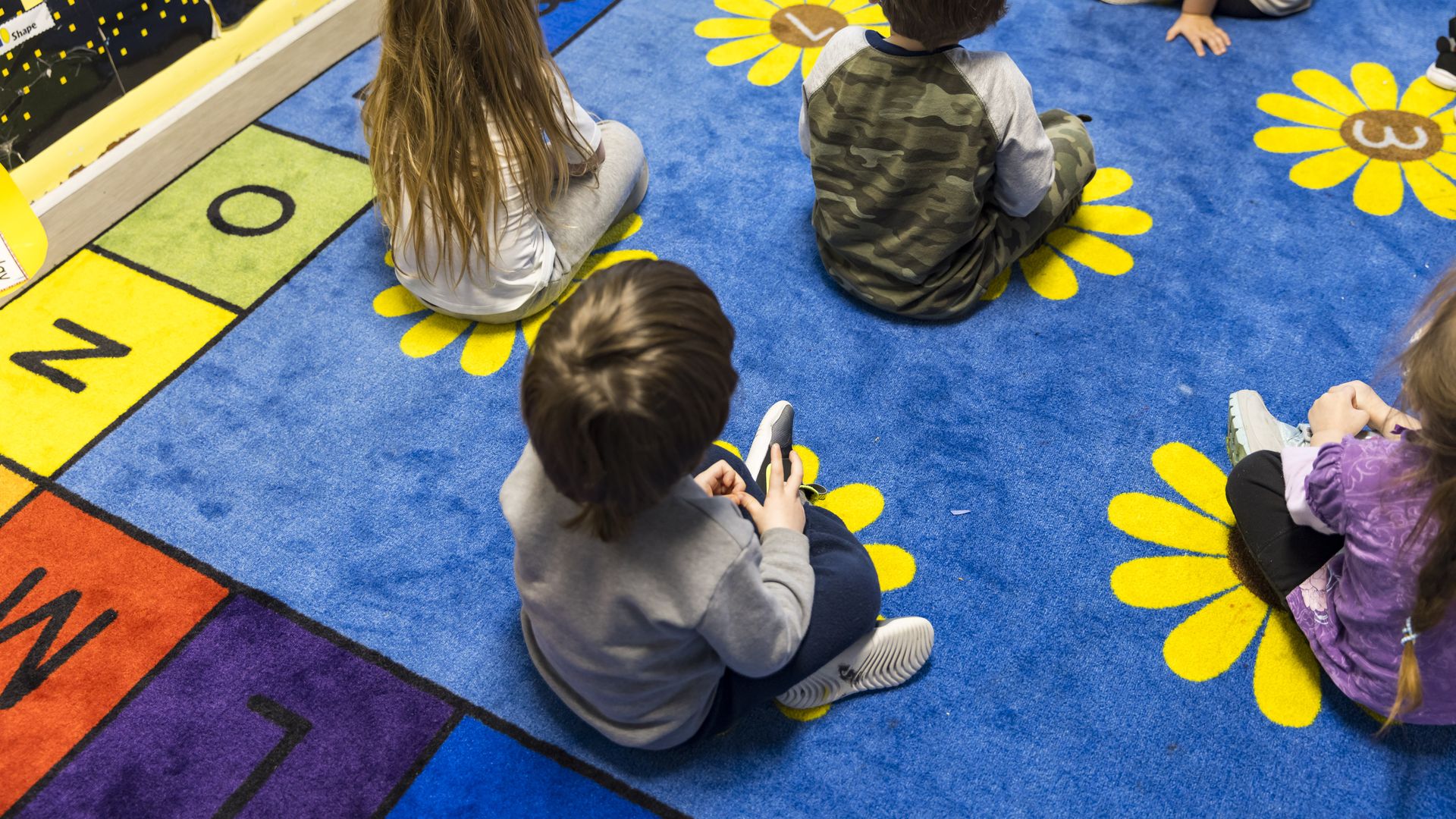 Children sit cross-legged on a multicolored rug in a classroom