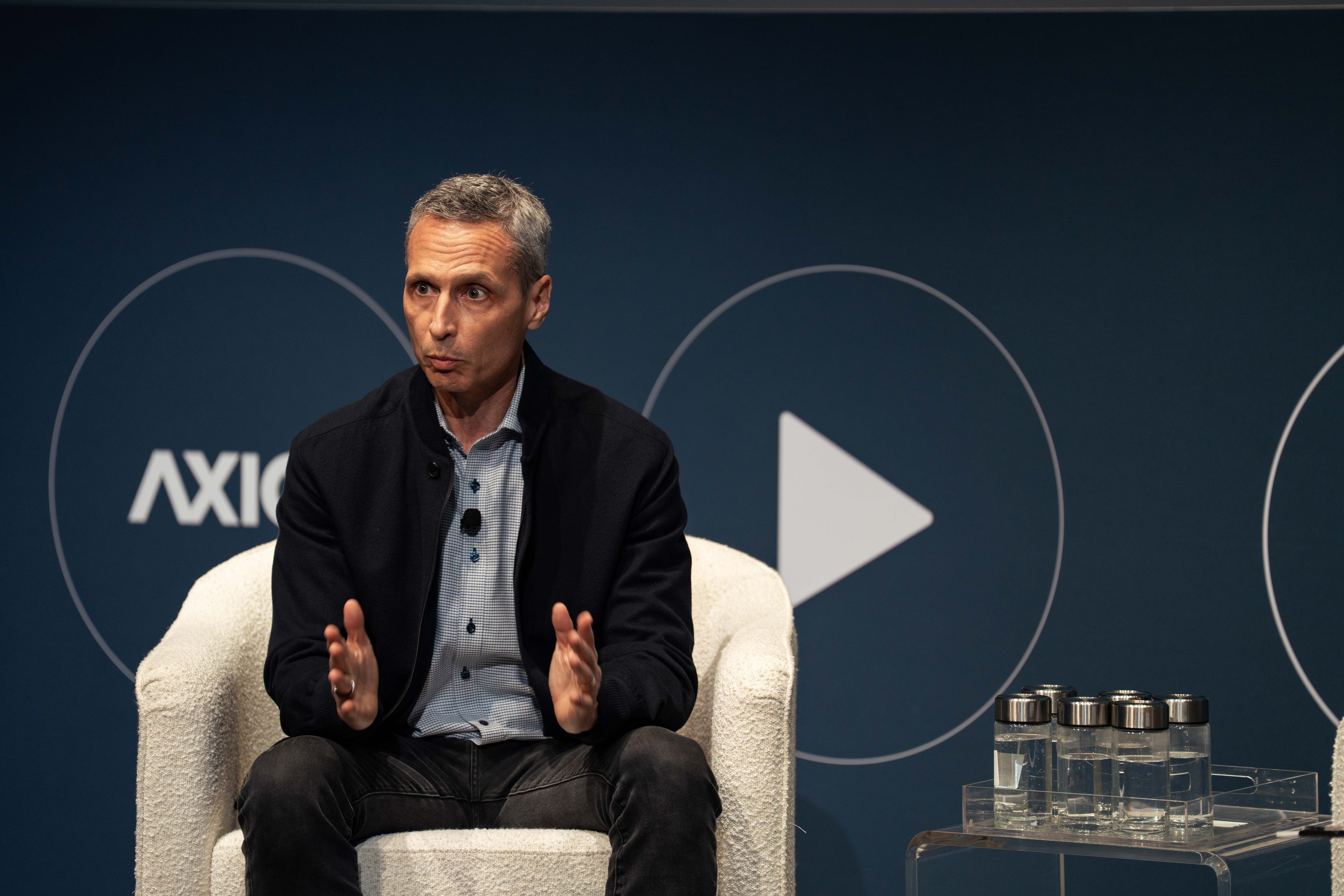 ESPN chair Jimmy Pitaro in a black jacket and checkered shirt speaking while sitting in a white chair on stage, dark blue background at Media Trends Live.