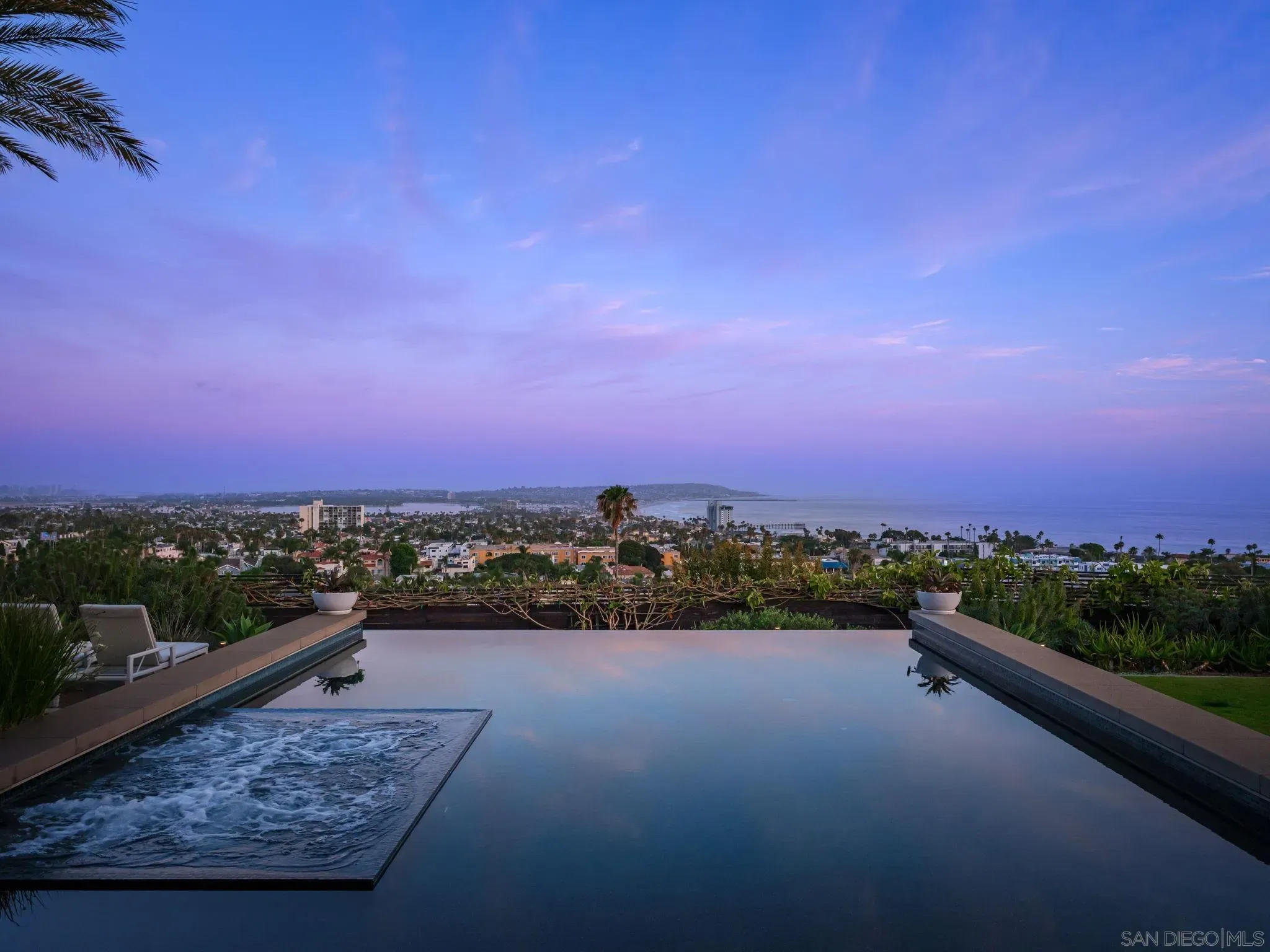 Twilight view of an infinity pool reflecting a purple-blue sky, with a palm frond at the left, lounge chairs on the deck, and a distant coastal city and sea on the horizon.