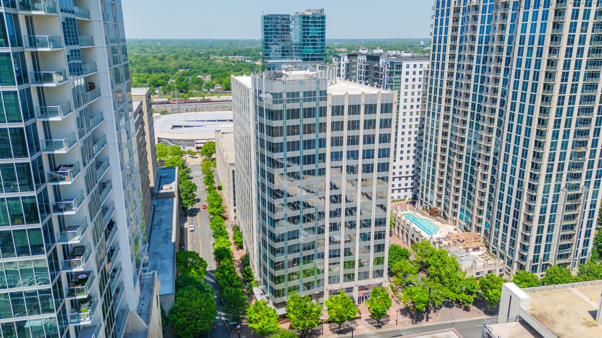 Aerial view of a city block with glass and concrete high-rise buildings, green trees lining streets, and a rooftop pool visible on one building under clear blue sky.