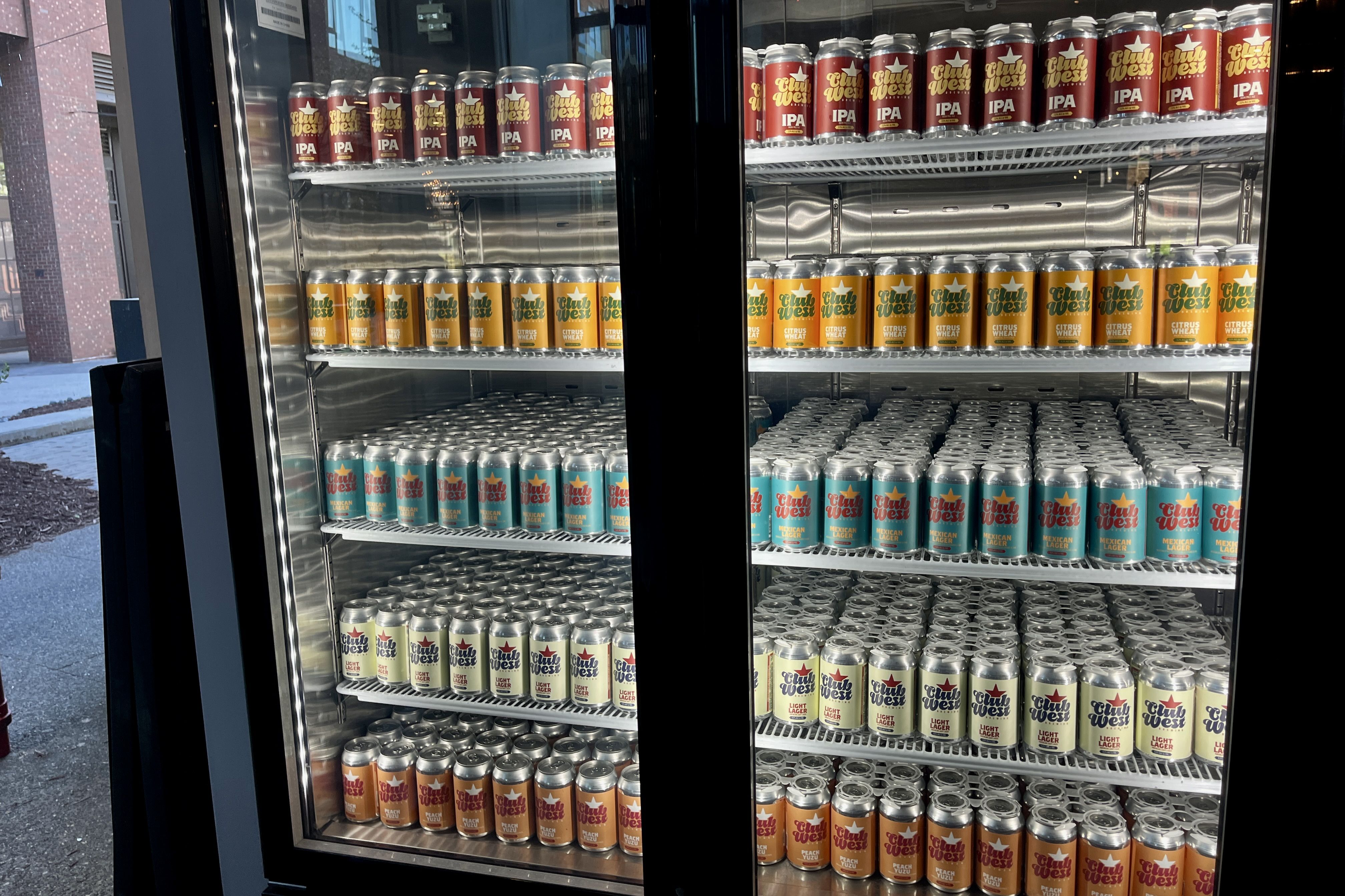 Glass-door refrigerator full of neatly arranged beer cans in red, orange, teal, and white labels on metal shelves, split by a black vertical divider in the middle.