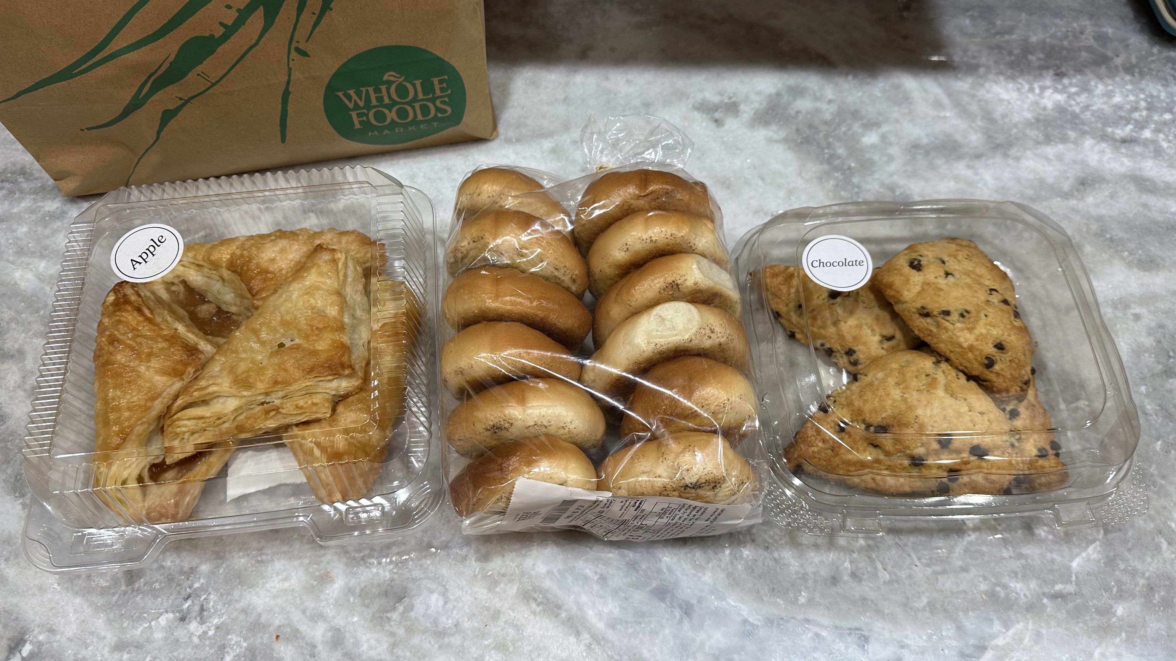 A box of apple turnovers, a bag of mini bagels and a box of chocolate chip scone sit on a counter next to a Whole Foods bag.