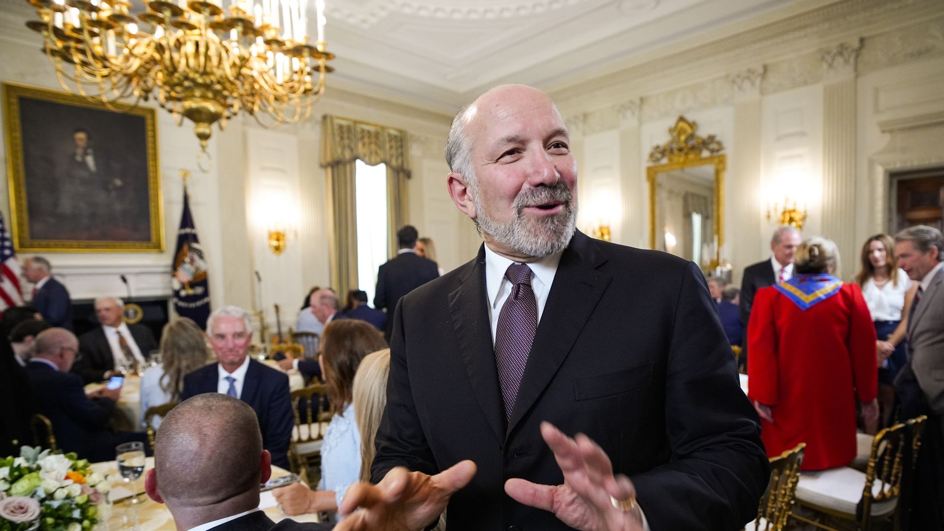 Howard Lutnick, in black suit and purple tie, gestures and smiles in an elegant room with chandelier, golden chairs, and several seated people in conversation during a formal event.