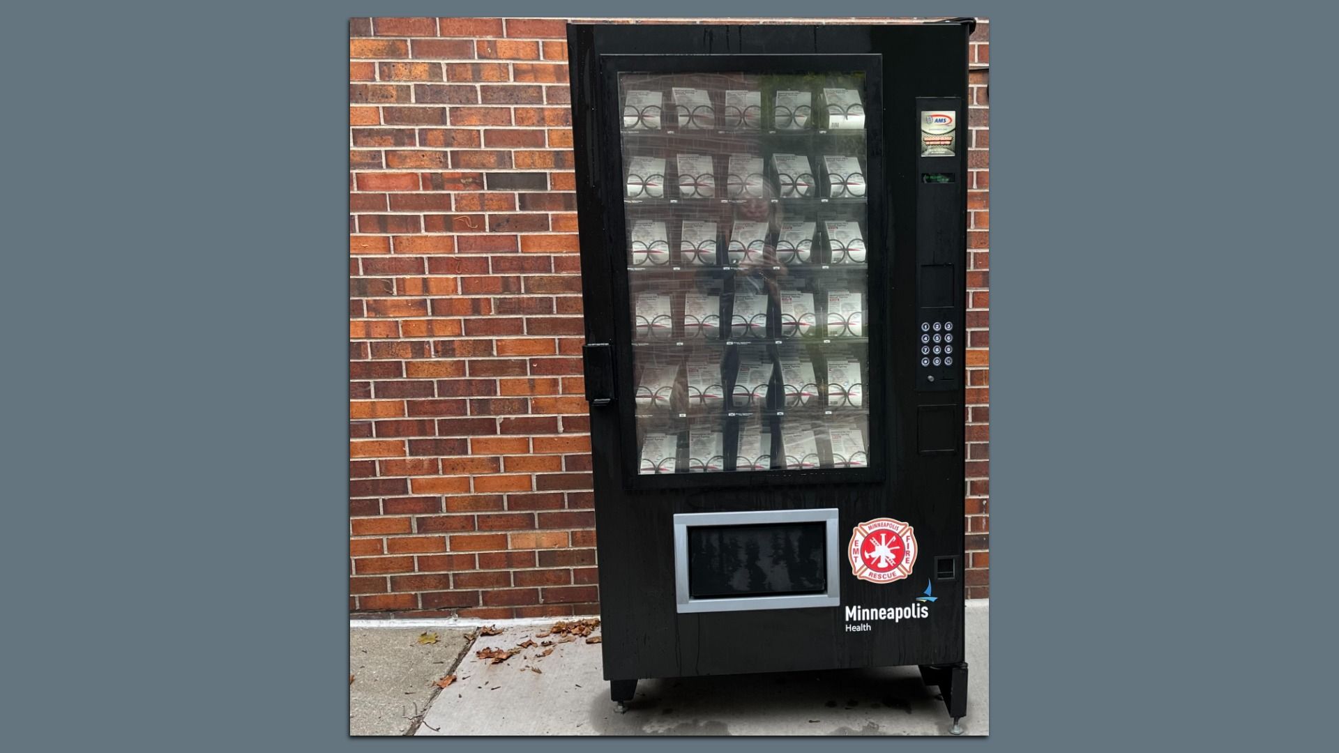 A black vending machine against a red brick wall, filled with white package items visible through the glass front. A keypad sits on the right side, and a Minneapolis Health sticker is near the bottom.