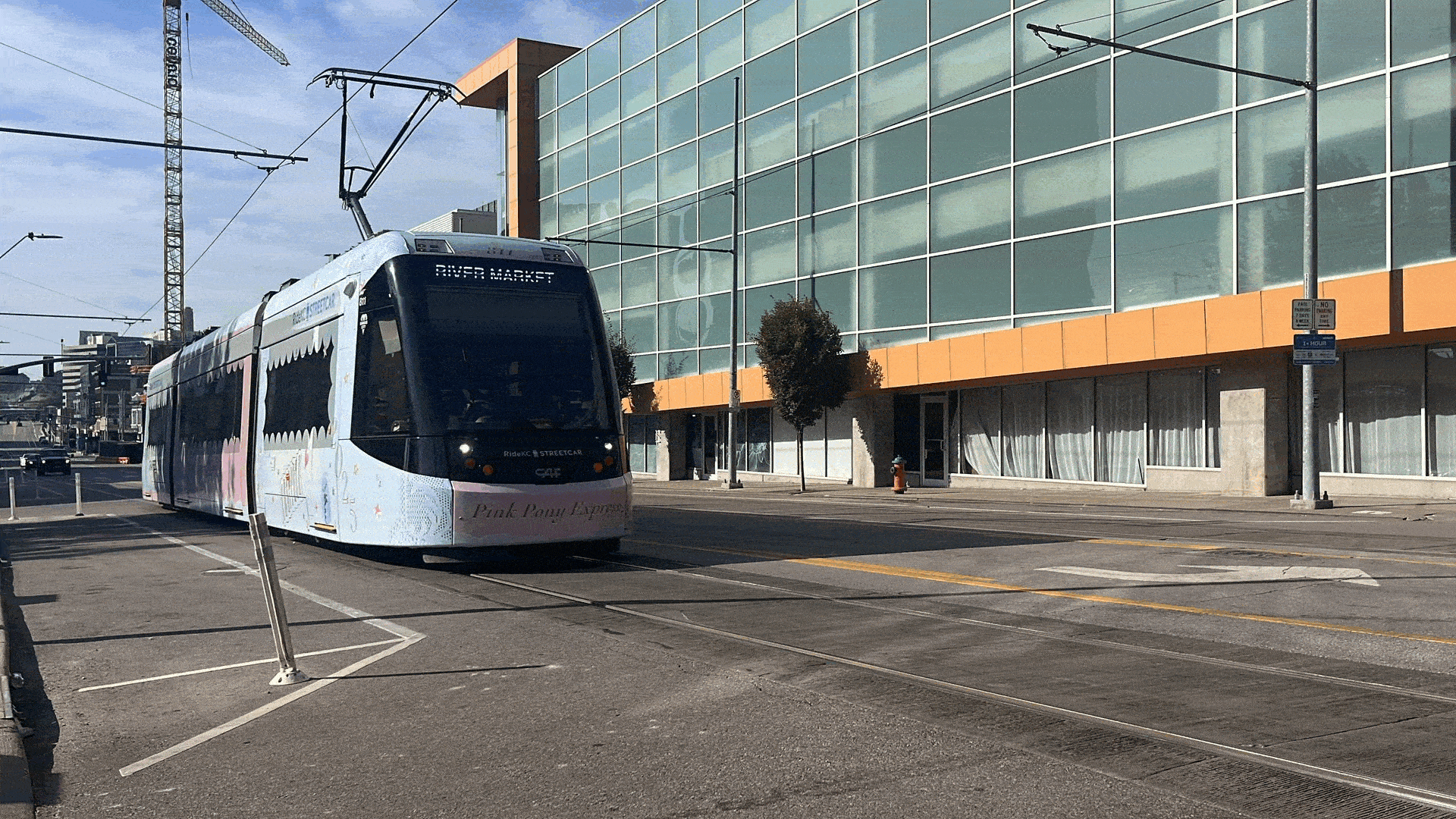 A pink and white streetcar labeled "Pink Pony Express" travels on tracks beside a large glass building with orange trim under a blue sky in an urban setting.