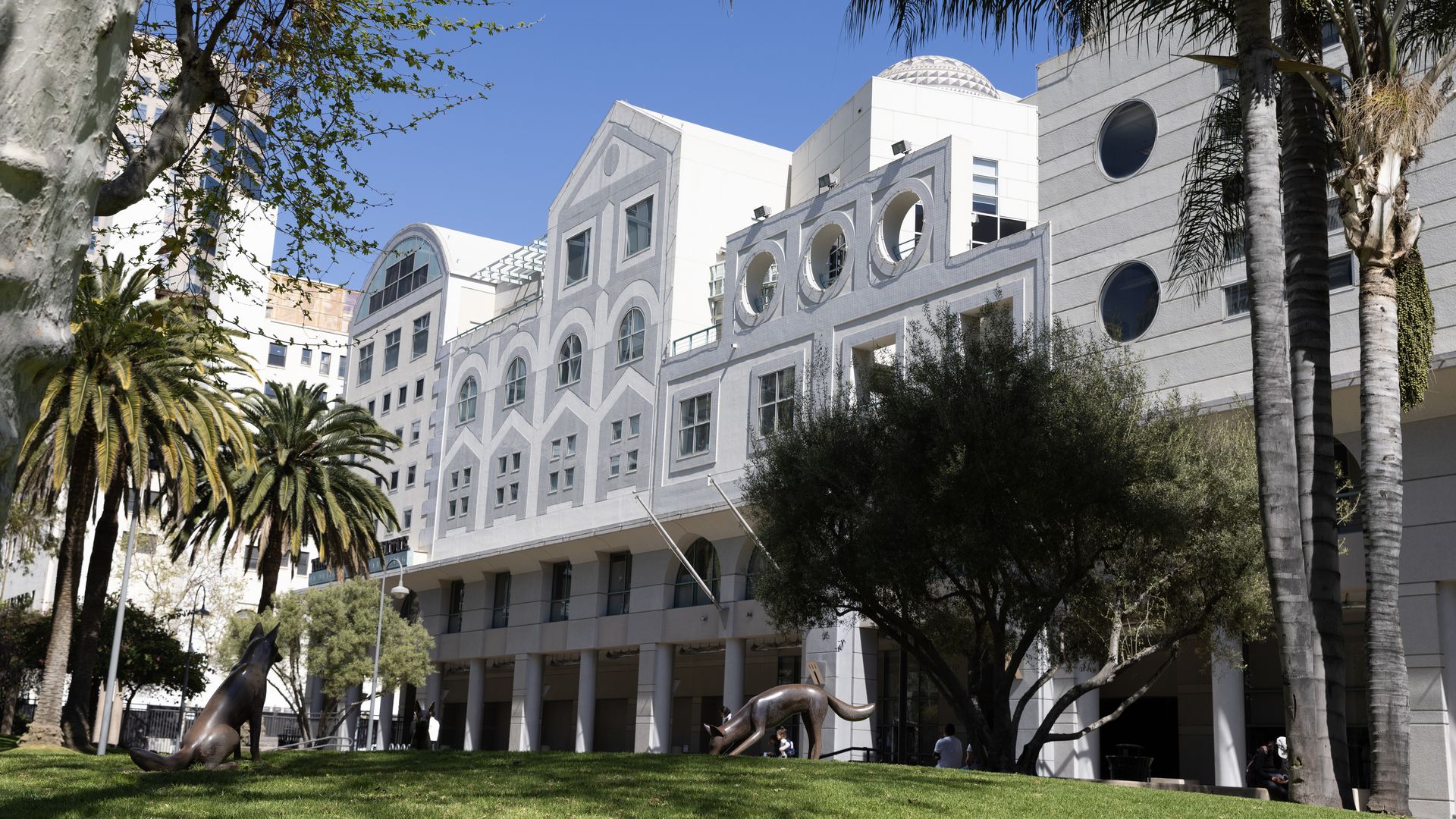 The facade of a white building with two metal dog sculptures and several palm trees in front. 