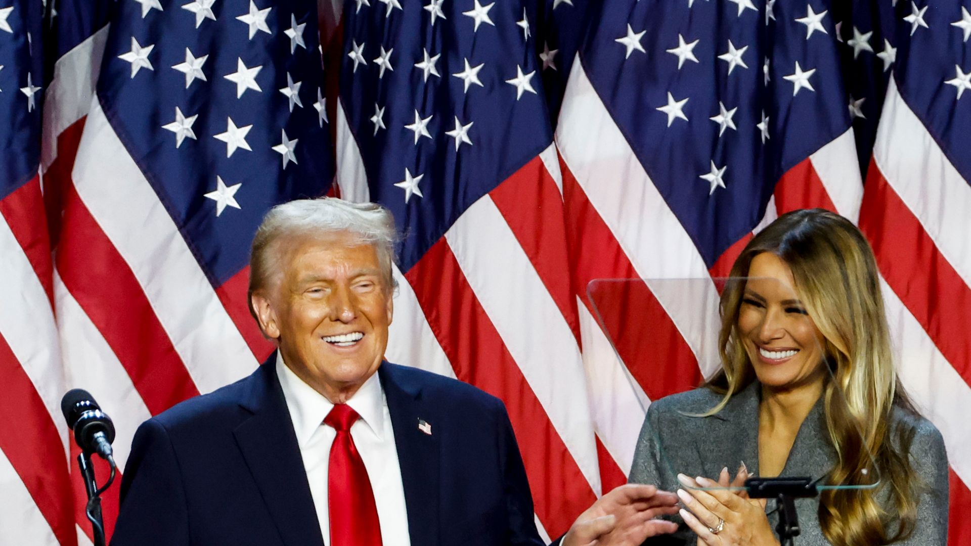 Donald and Melania Trump smile in front of several U.S. flags.