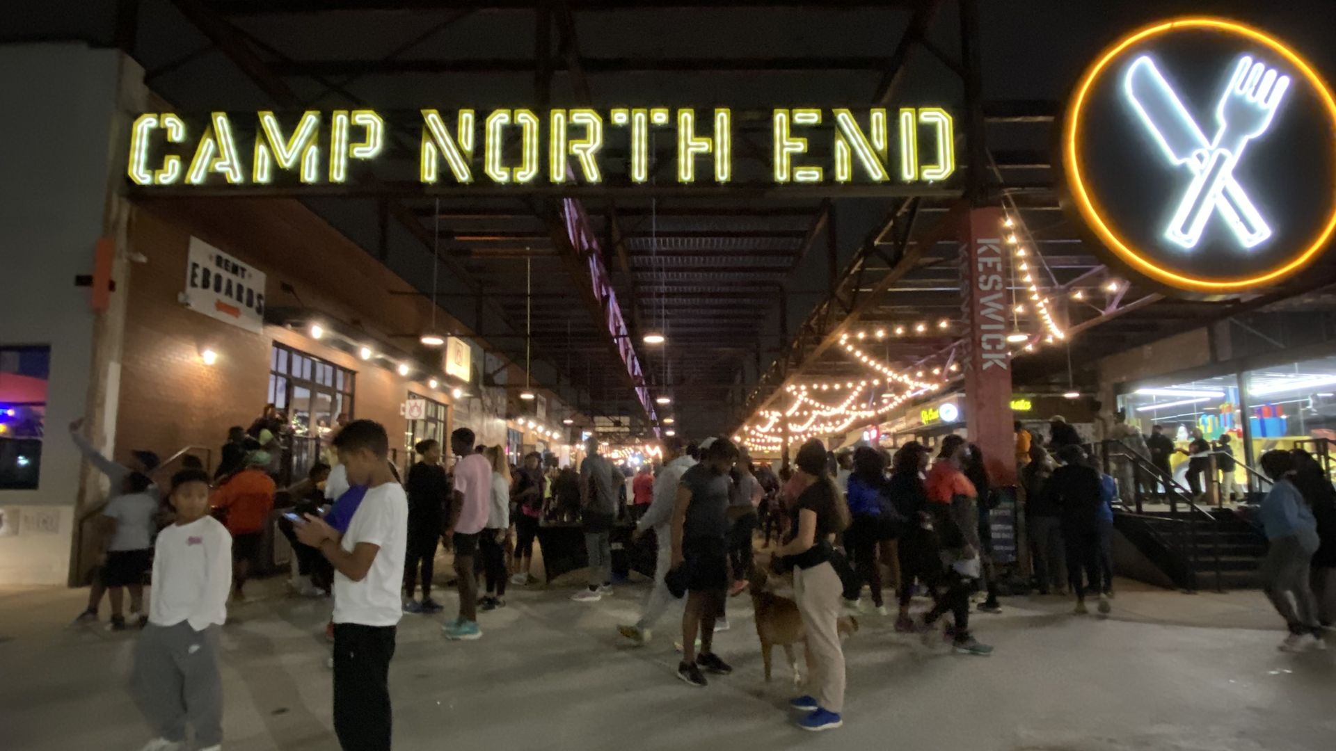 Nighttime crowd under a bright yellow neon sign reading "CAMP NORTH END", with string lights and a round neon sign showing a fork and knife crossed on the right.