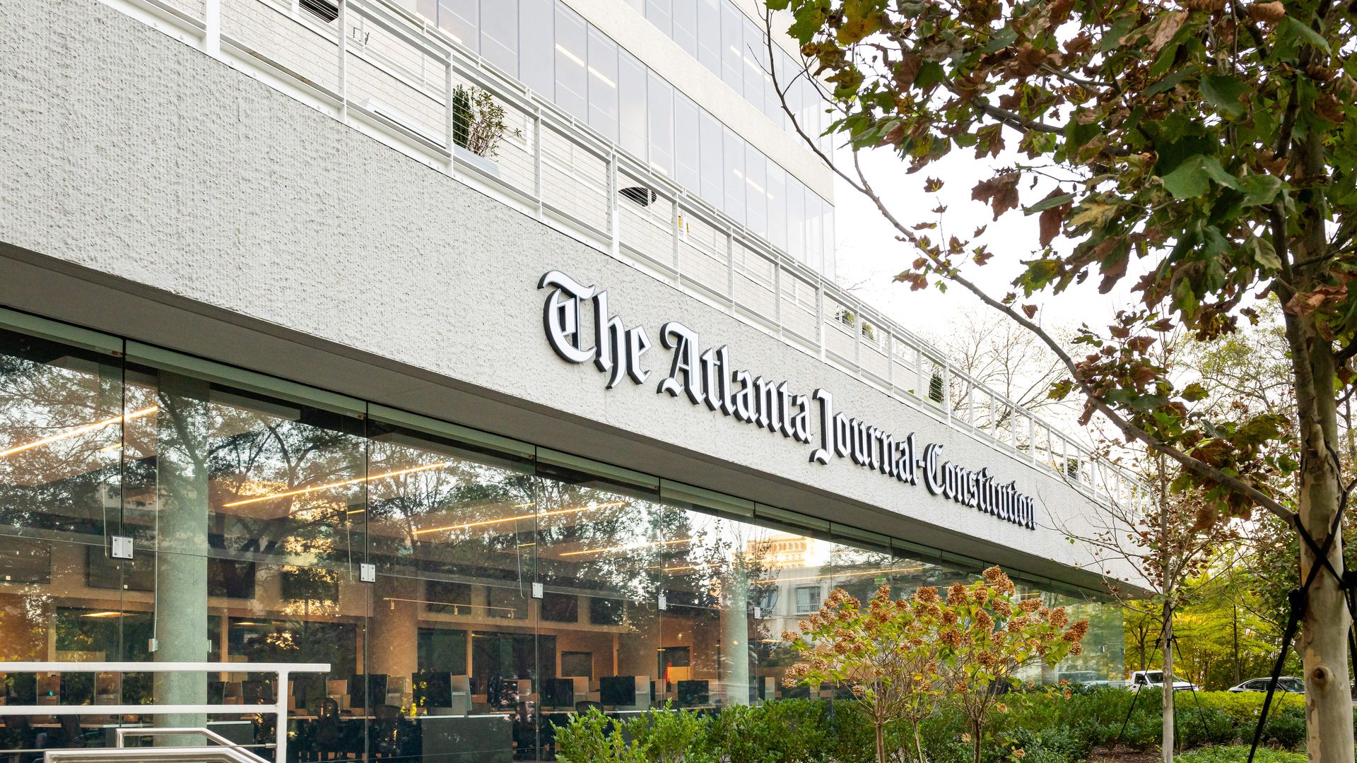 Exterior of The Atlanta Journal-Constitution building with glass windows reflecting trees and greenery, plants in front, and a tree with autumn leaves on the right.