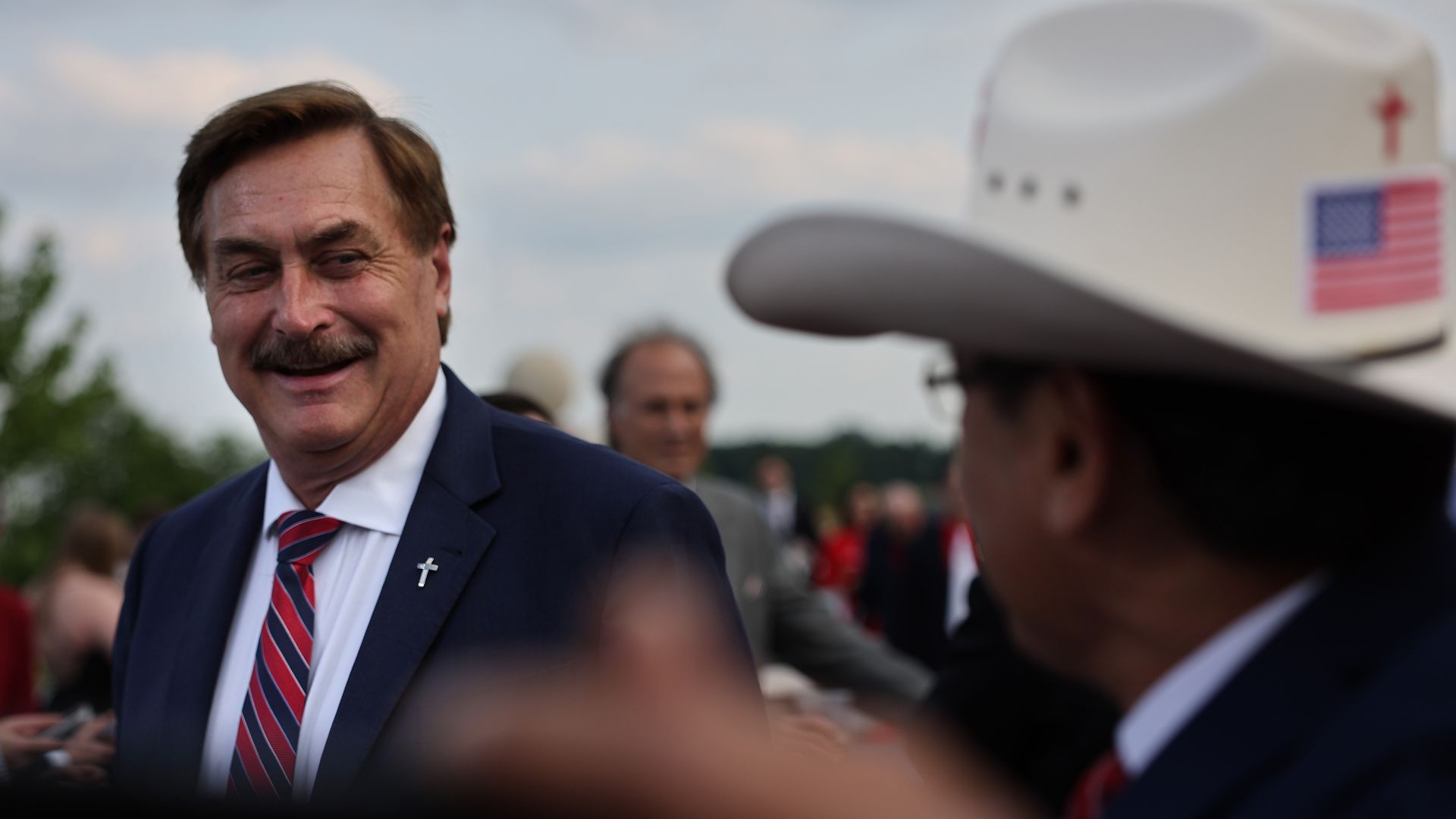 Mike Lindell wearing a blue suit and red/blue tie smiles at a man in a cowboy hat with an American flag and a cross on the back. Lindell's suit has a cross pin on it. 