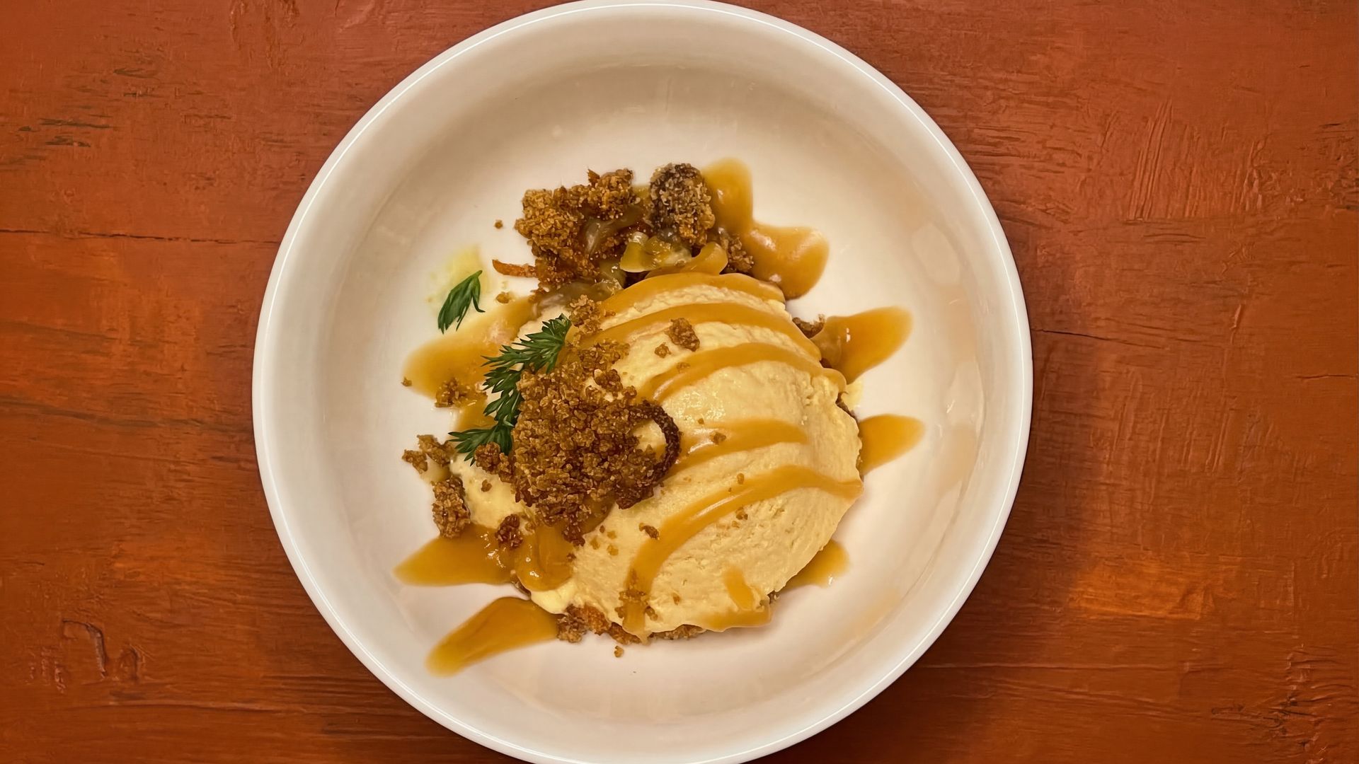 Top-down view of a white bowl with sliced chicken, drizzled sauce, crunchy brown granola topping, and a small sprig of green herb, on a reddish wooden table.