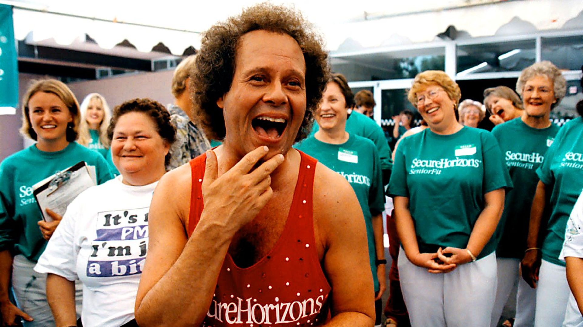 Richard Simmons, wearing a red "SecureHorizons SeniorFit" tank top, smiles and gestures with his hand at his chin while standing in front of a cheerful group of women in green "SecureHorizons SeniorFit" shirts. 