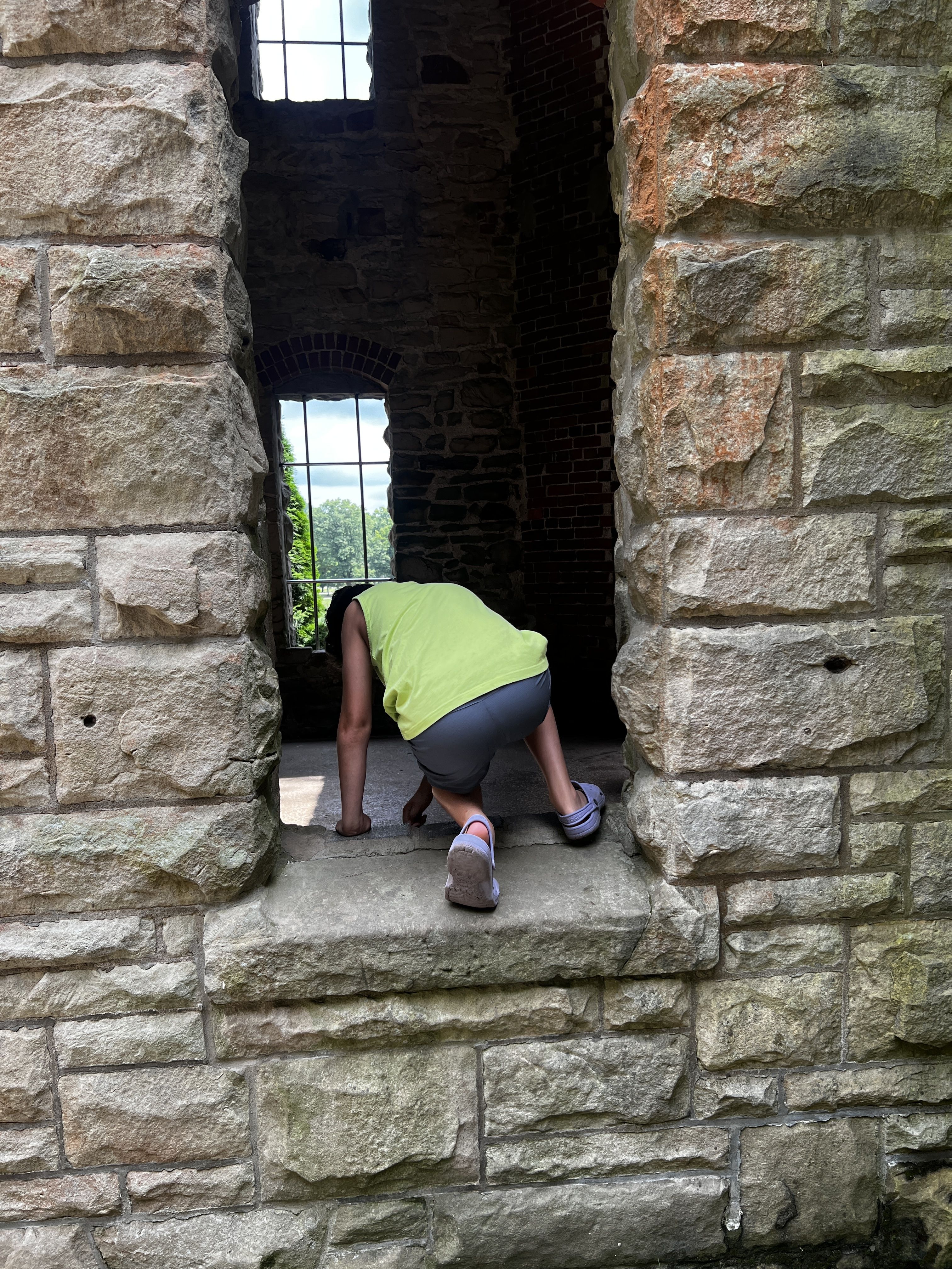 Person in a neon green shirt and gray shorts crouches on a stone ledge inside a ruined brick-and-stone structure, bending toward the ground near an arched window with bars; green trees visible outside.