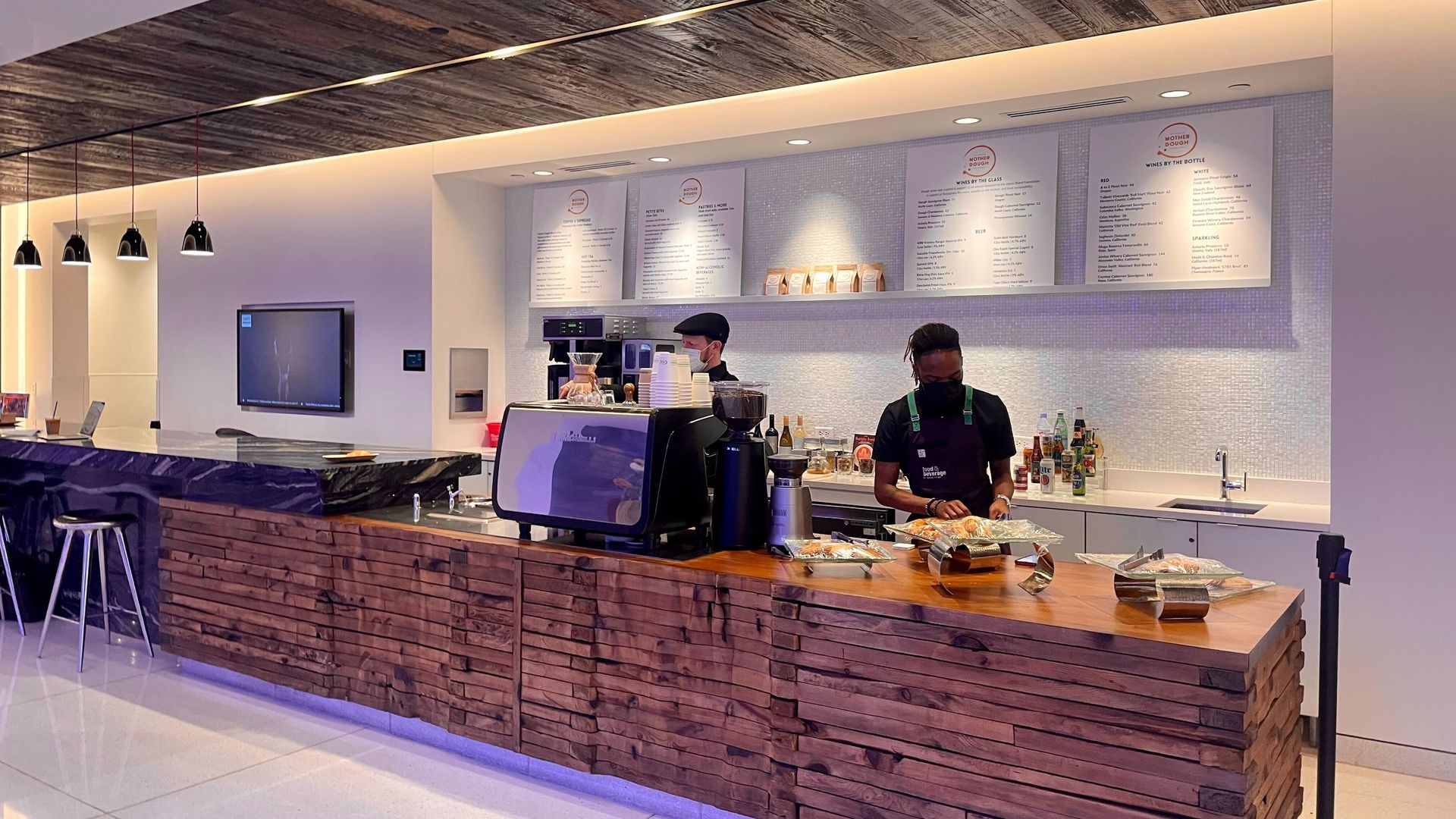 A long wooden countertop with two men standing behind it. A menu and pastries are seen in the background.