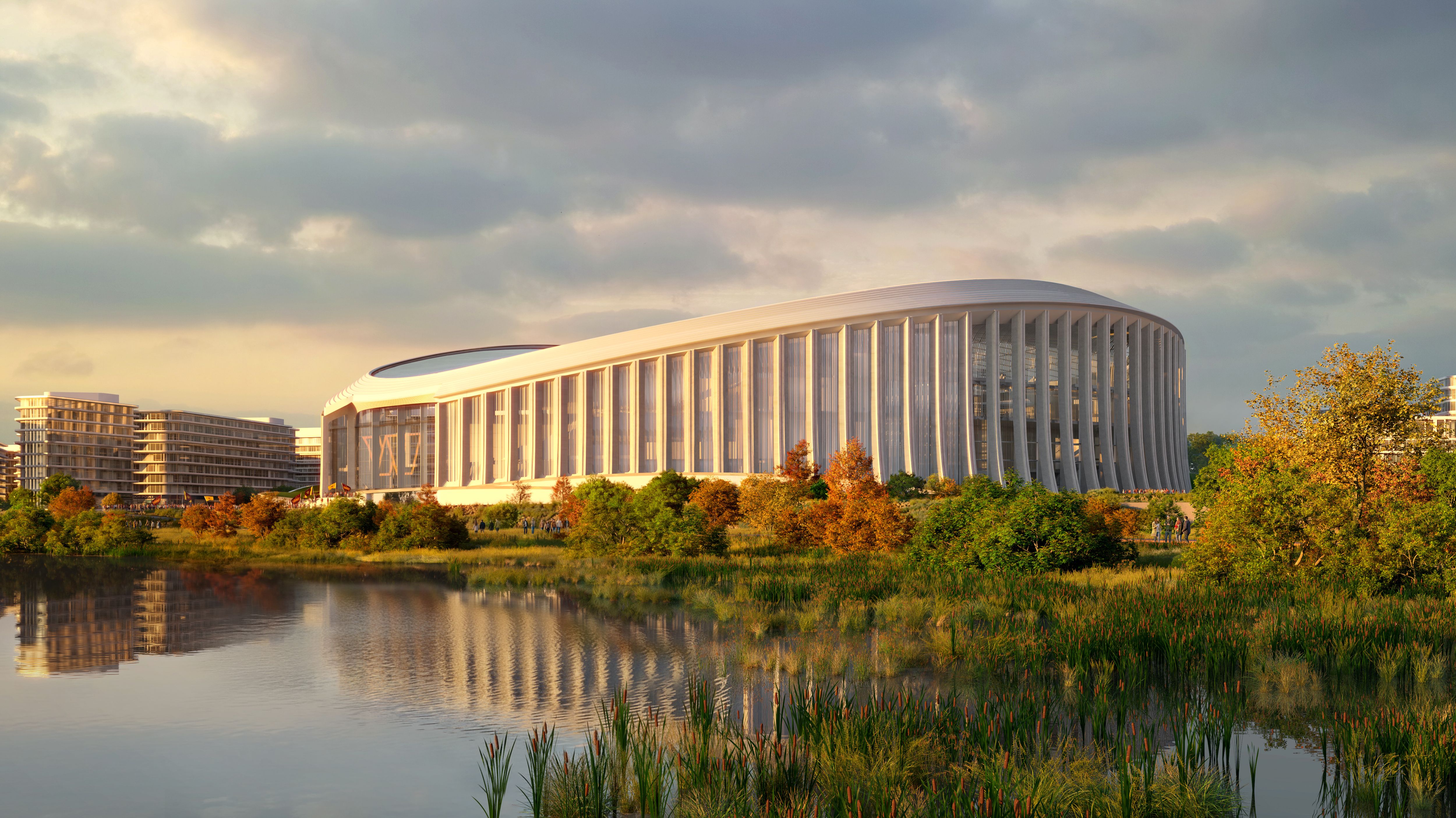 Modern oval building with vertical white columns by a pond, surrounded by green and autumn-colored trees, under a partly cloudy sky at sunset.