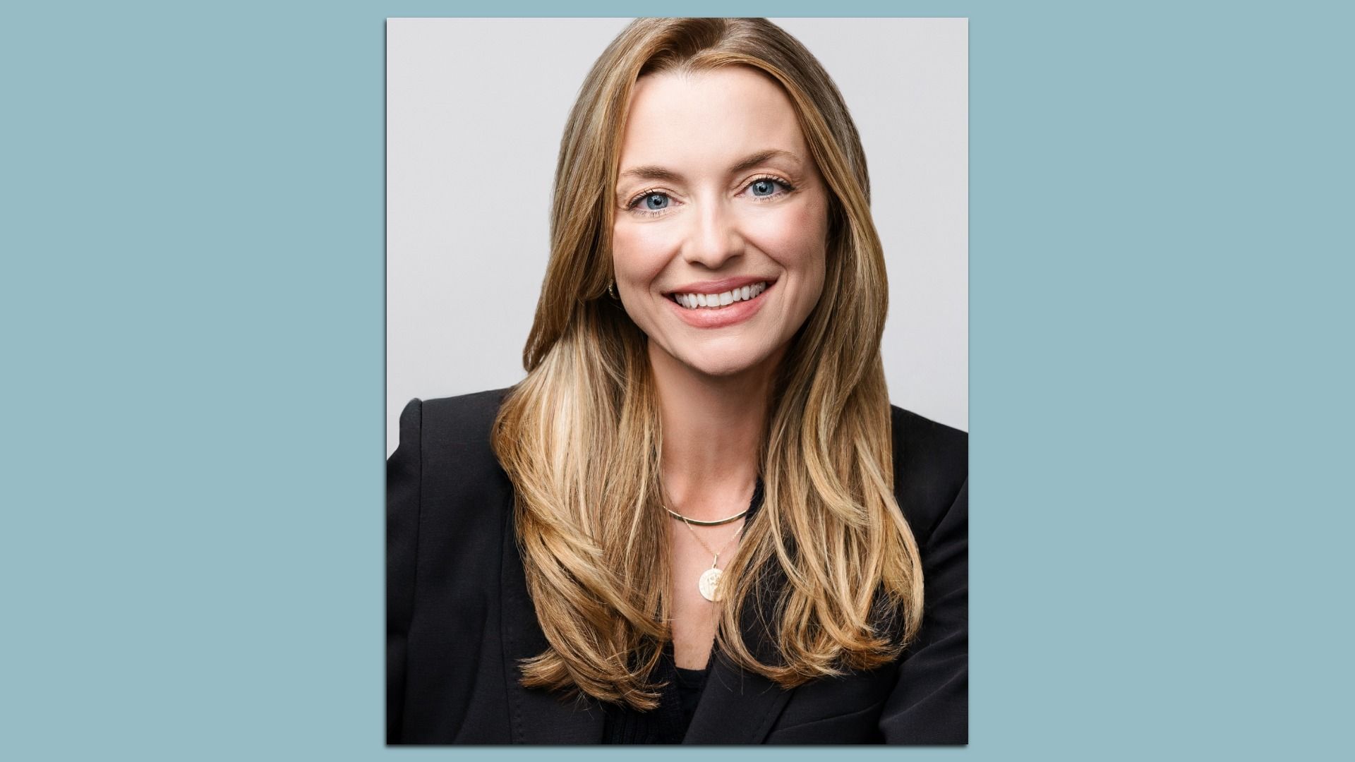 Studio portrait of a smiling blonde woman in a black blazer with a gold necklace, against a light gray backdrop.