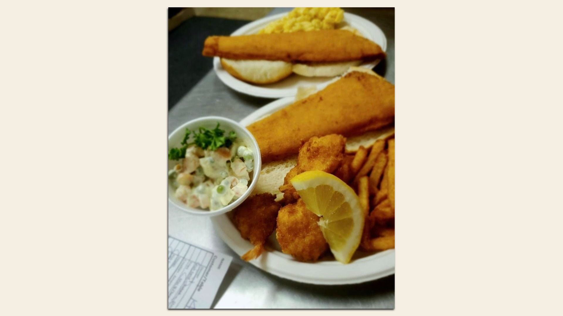 Golden fried fish with French fries and a lemon wedge on a plate; side of tartar sauce with parsley. A long fried item rests on a second plate in the background.