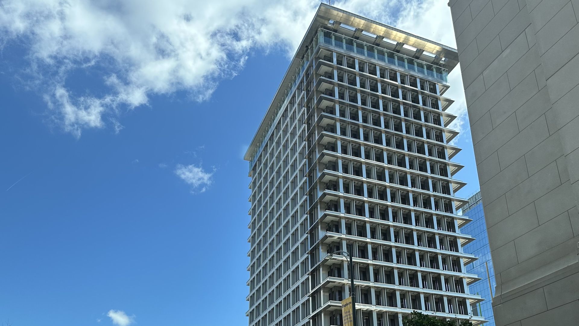 Low-angle view of a tall, modern glass-and-grid apartment building with rows of balconies, set against a bright blue sky with scattered clouds; a beige wall on the right and trees at the bottom.