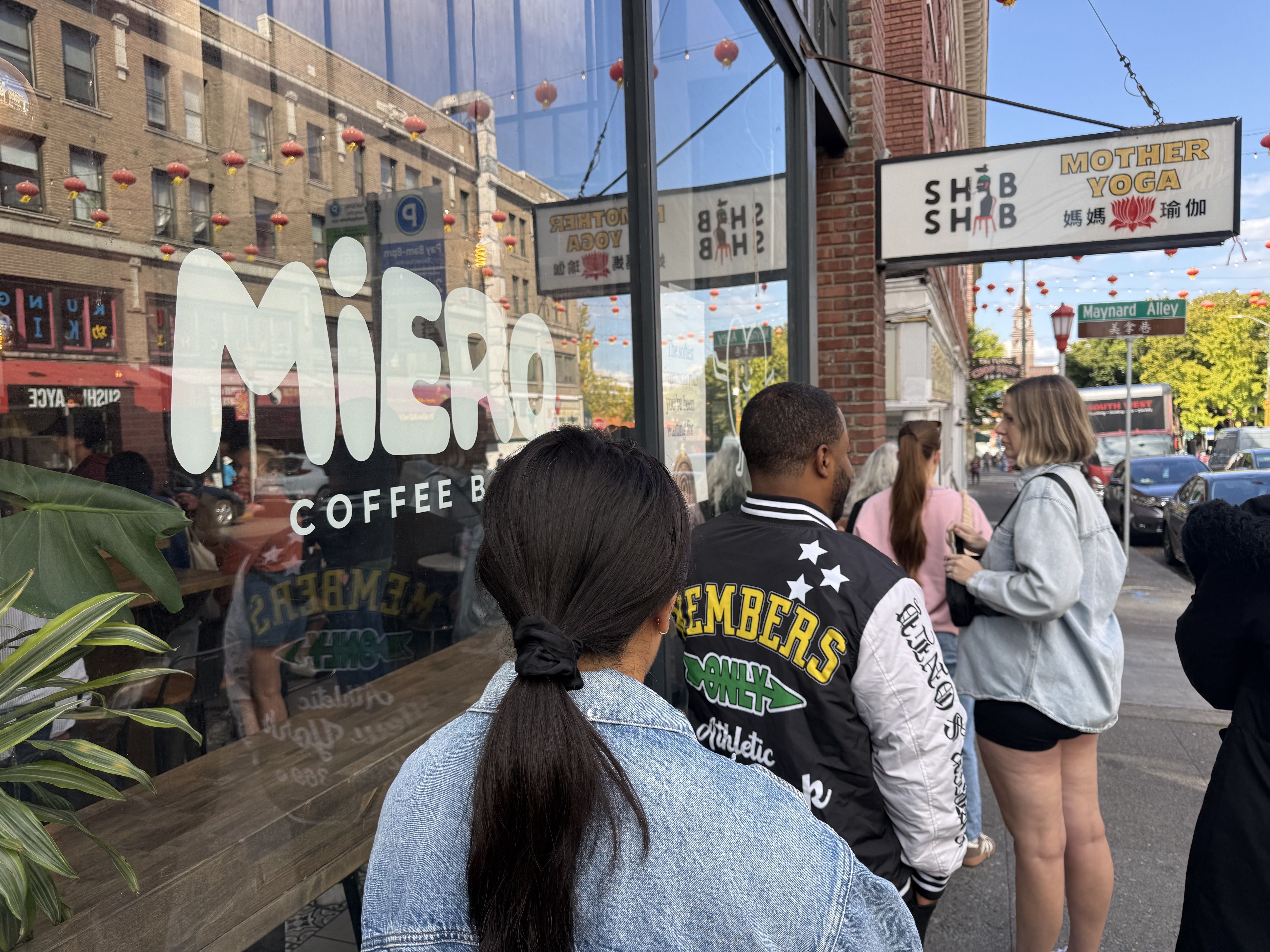 Busy city street outside a cafe; a large glass window displays white letters. Red lanterns hang overhead. A brick storefront sign reads "MOTHER YOGA". People in jackets line up along the sidewalk.