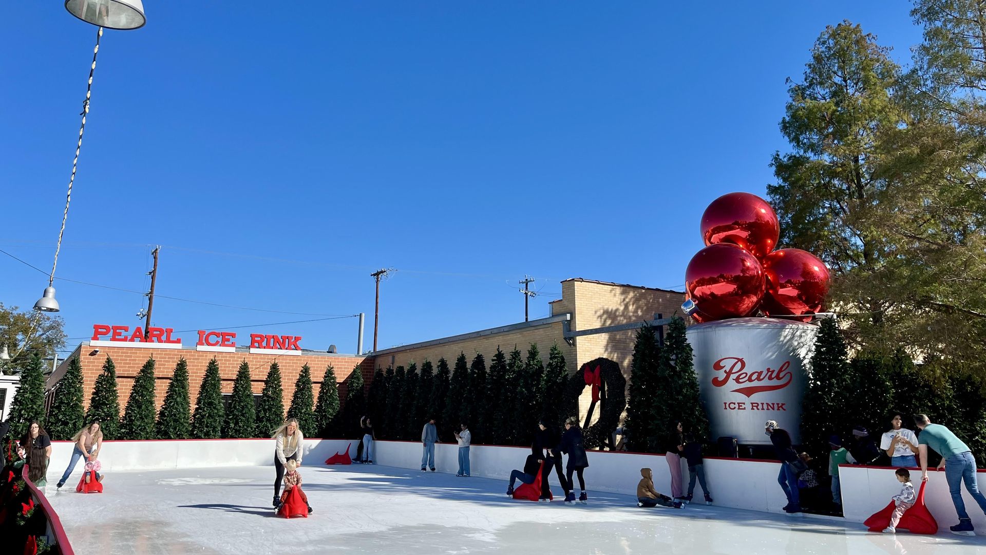 An outdoor ice rink with people skating on red skating aids. A large silver mug decoration that reads "Pearl" is seen in the background with three big red ornaments on top of it. A sign reads "Pearl ice rink."