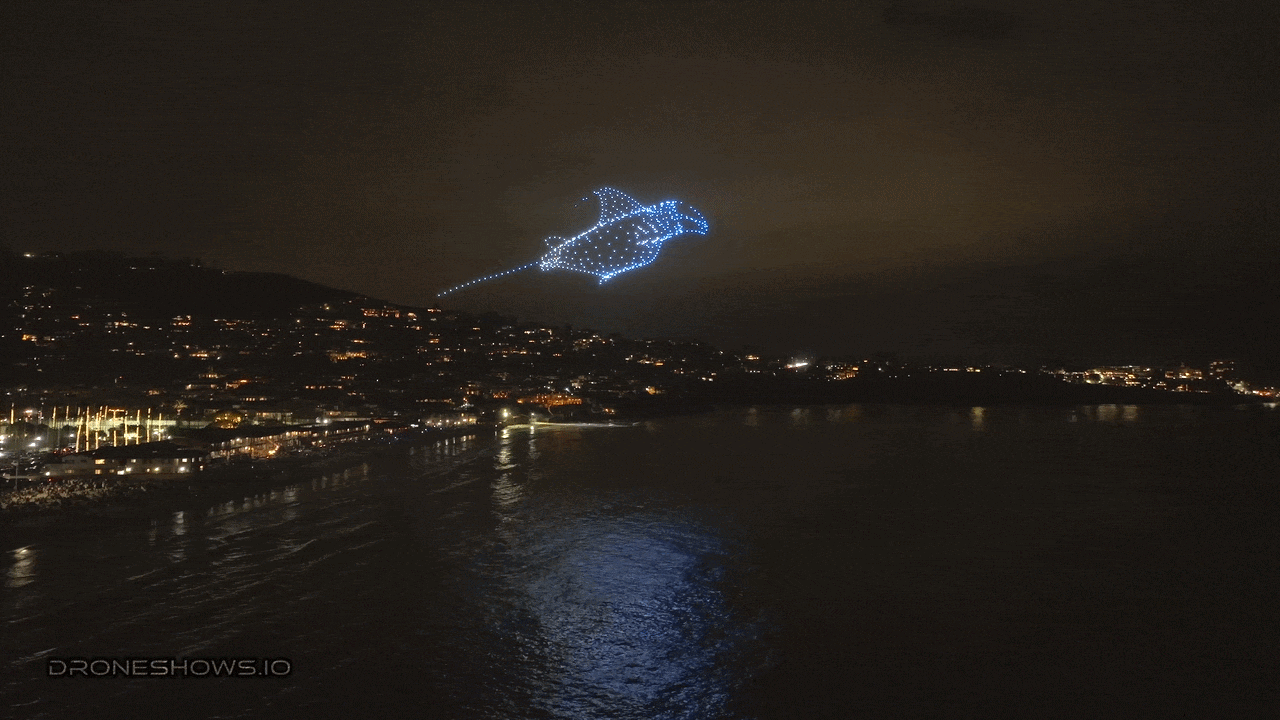 Night aerial view of a coastal city with warm shoreline lights; blue drone-light formation in the sky shapes a manta ray, with a glowing trail over the water.