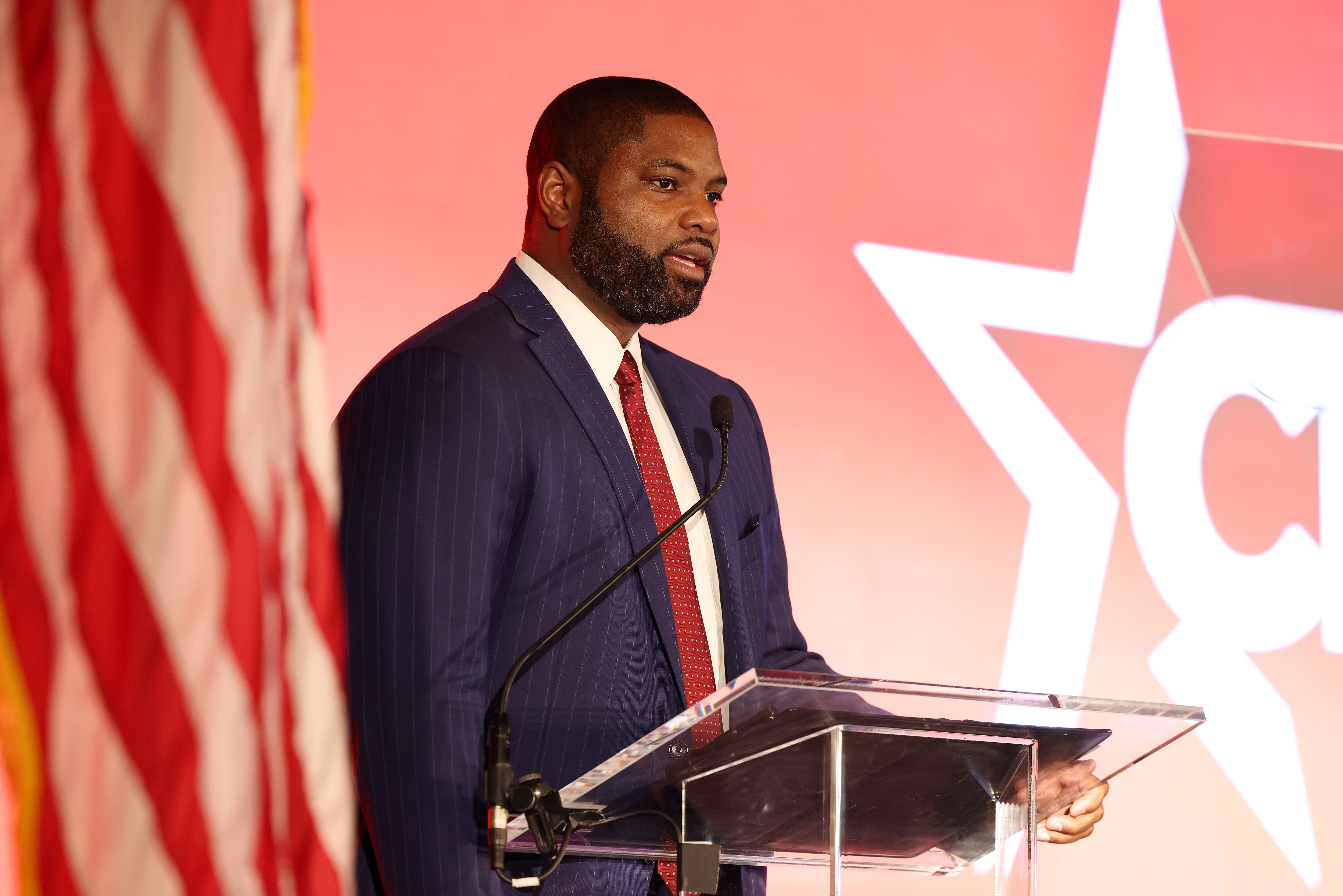 Rep. Byron Donalds speaks at a podium with a microphone, standing in front of a large graphic star on a red background. An American flag is visible on the left side of the image.