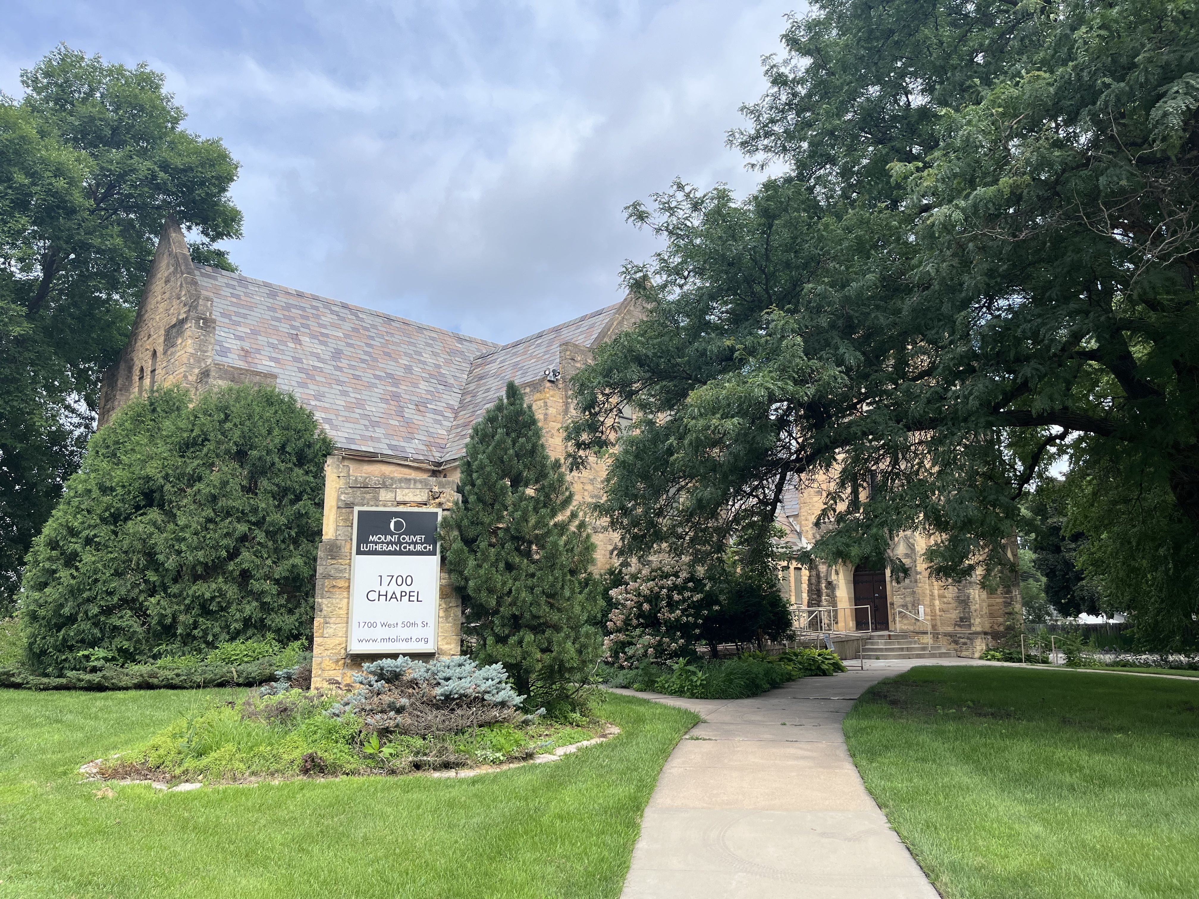 Stone building of Mount Olivet Lutheran Church surrounded by green trees, bushes, and grass. A curved sidewalk leads to the chapel entrance under a cloudy sky.