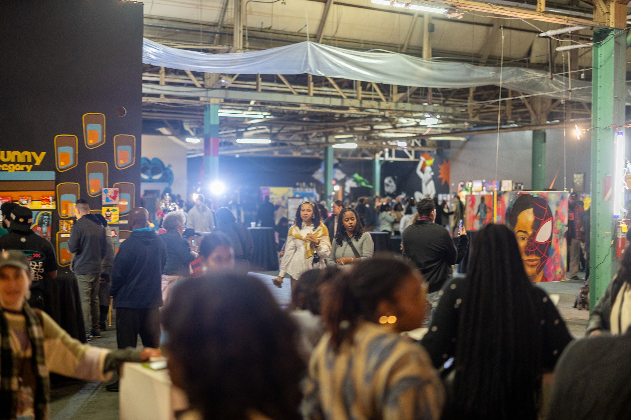 Indoor art exhibit with diverse crowd, colorful paintings, and murals including a split face with Spider-Man mask, under high industrial ceiling with beams and hanging fabric.