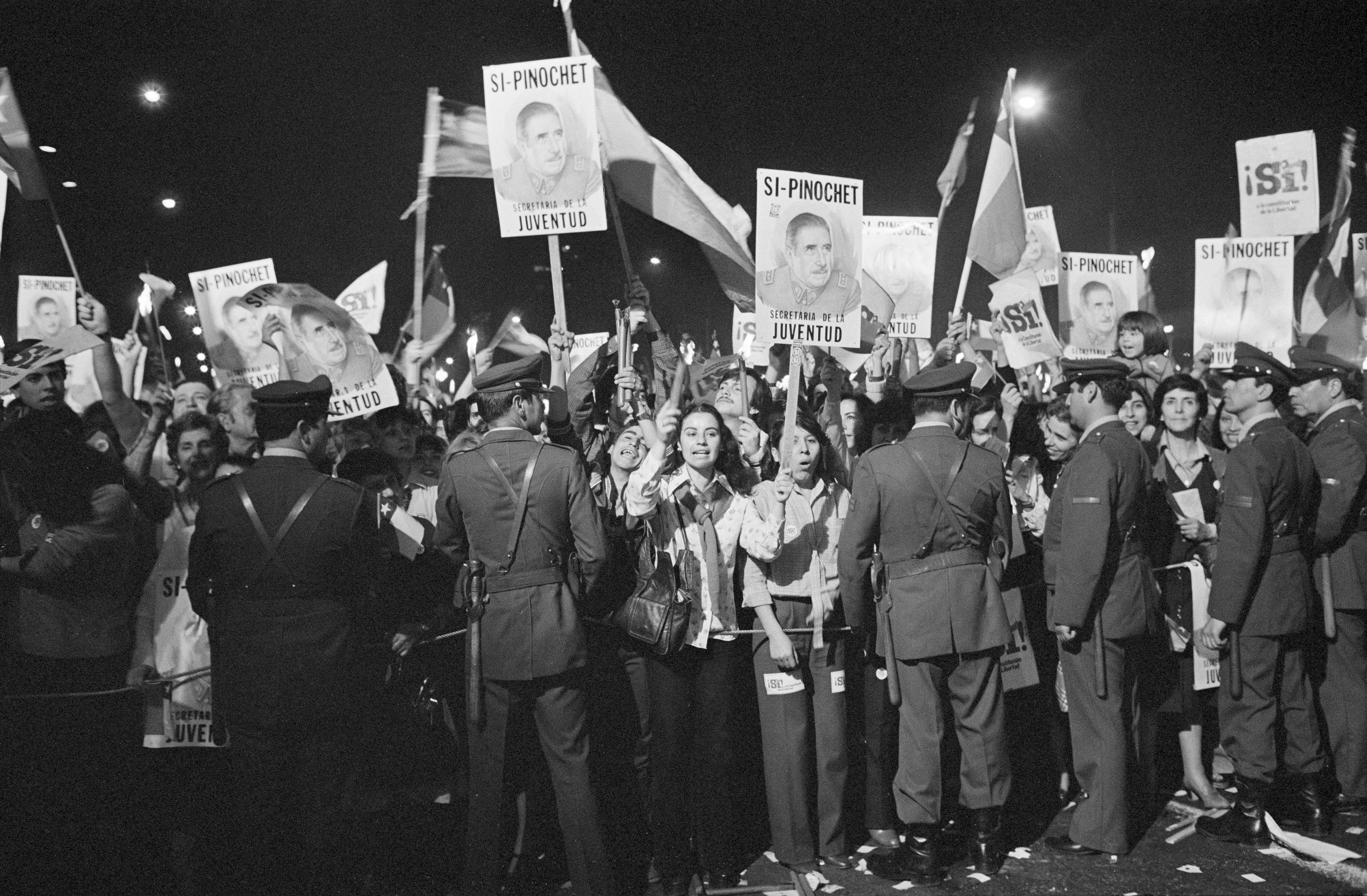 Protesters hold white signs with the face of Augusto Pinochet in this black and white photo. Police, with their backs to the camera, hold the line back. 