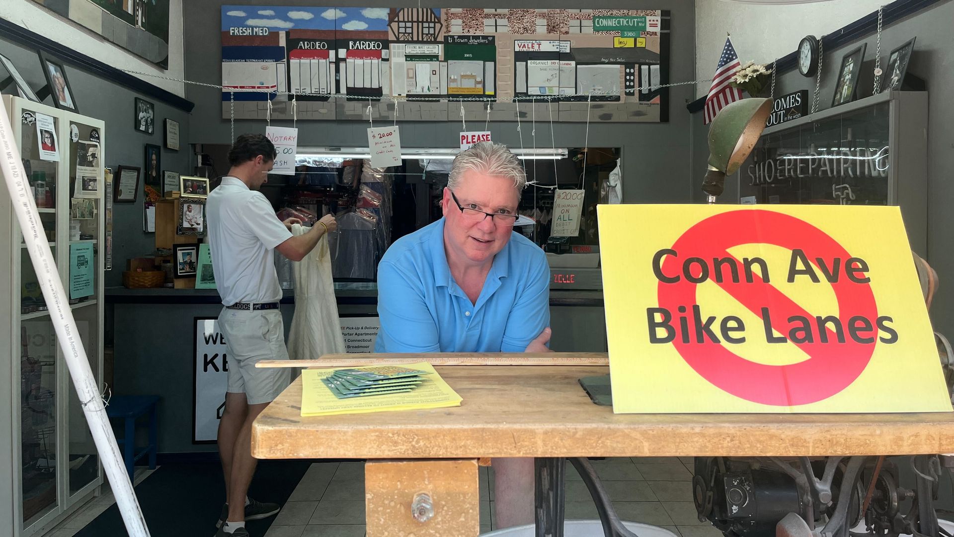Dry cleaner shop owner poses next to anti "Conn Ave Bike Lanes" sign on storefront