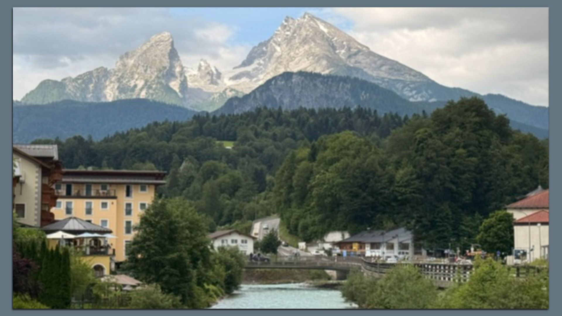 Snowy mountains rise above a dense forest; in the foreground a river runs through a small town with yellow and beige buildings, greenery, and a wooden railing along the bank.