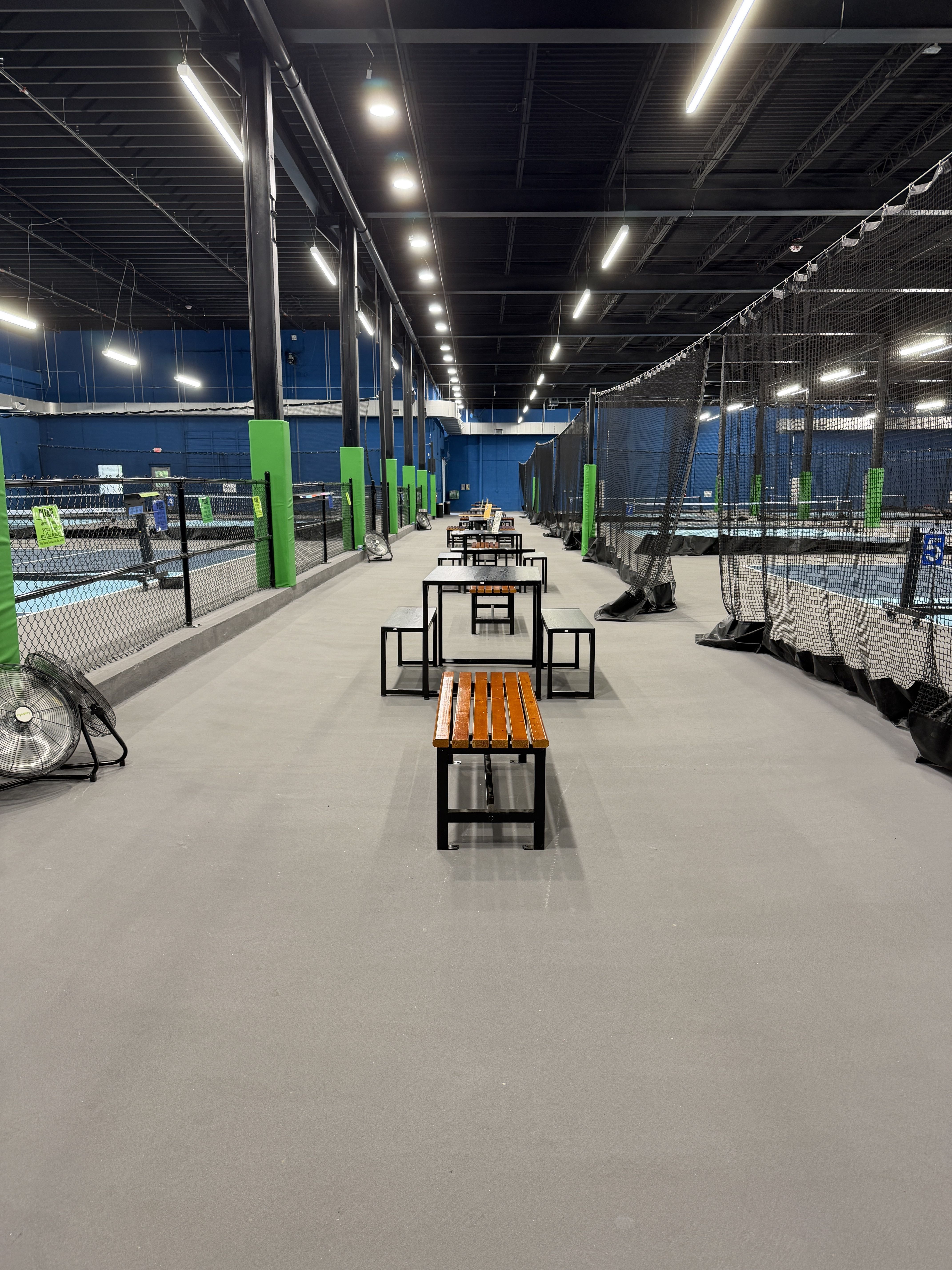 Indoor sports facility with a high dark ceiling and bright overhead lights. Blue walls, green padded posts, and netted cages along the right; benches line the central aisle.