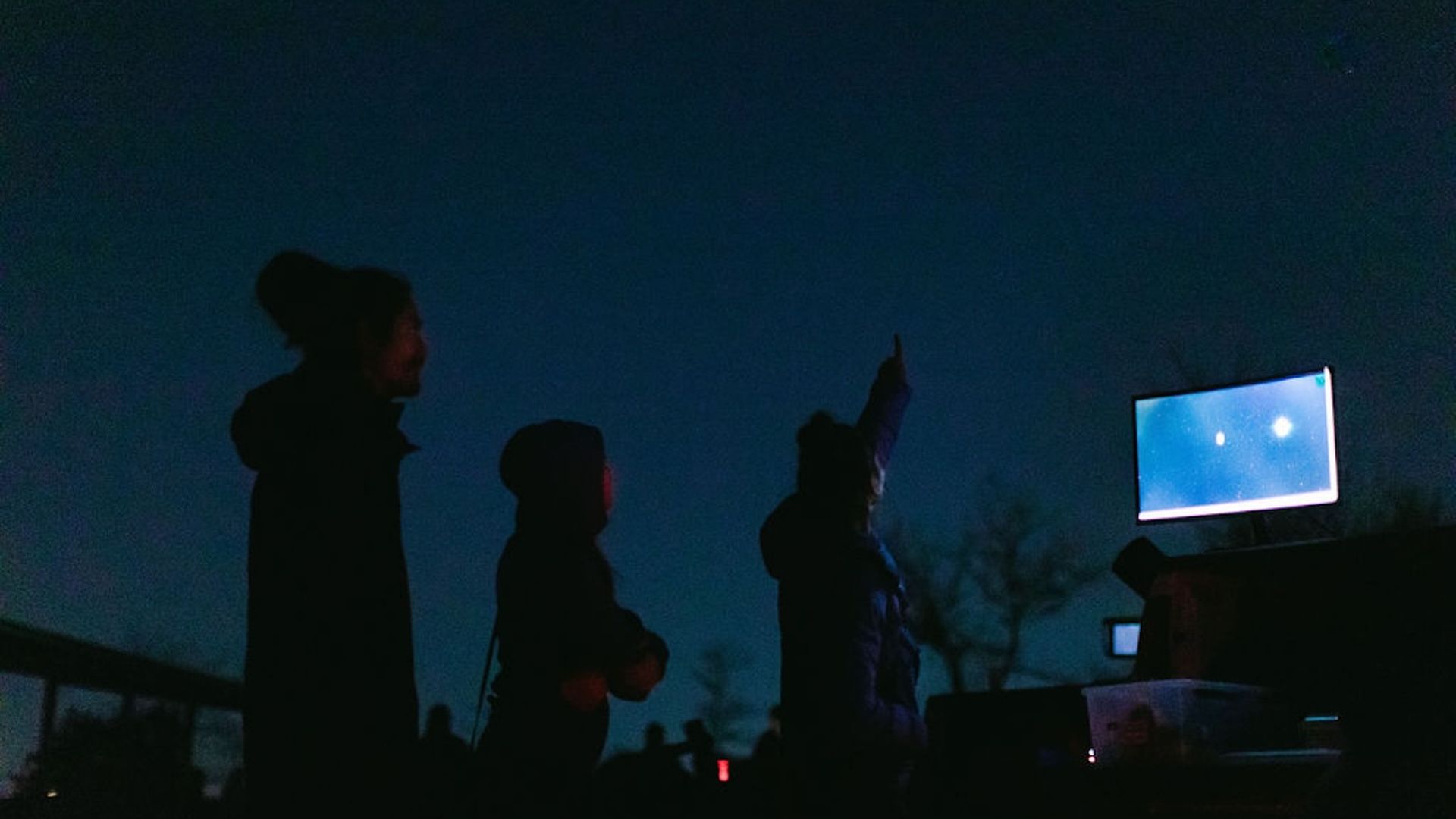 Silhouetted group outdoors at dusk, one person raises an arm toward a glowing blue screen on a monitor to the right, while others watch in dark jackets under a deep blue night sky.