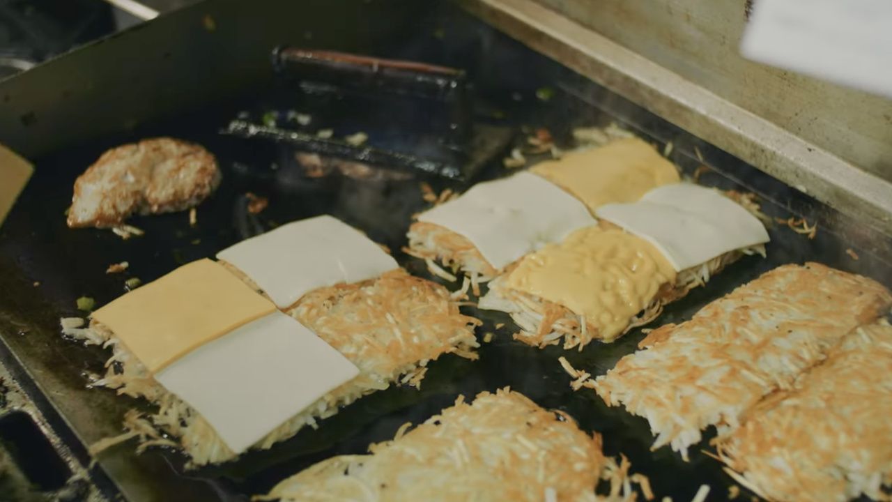 On a flat-top griddle, several hash-brown patties with shredded potato edges. Some are topped with white cheese slices, others with a yellow sauce; a lone browned patty sits nearby.
