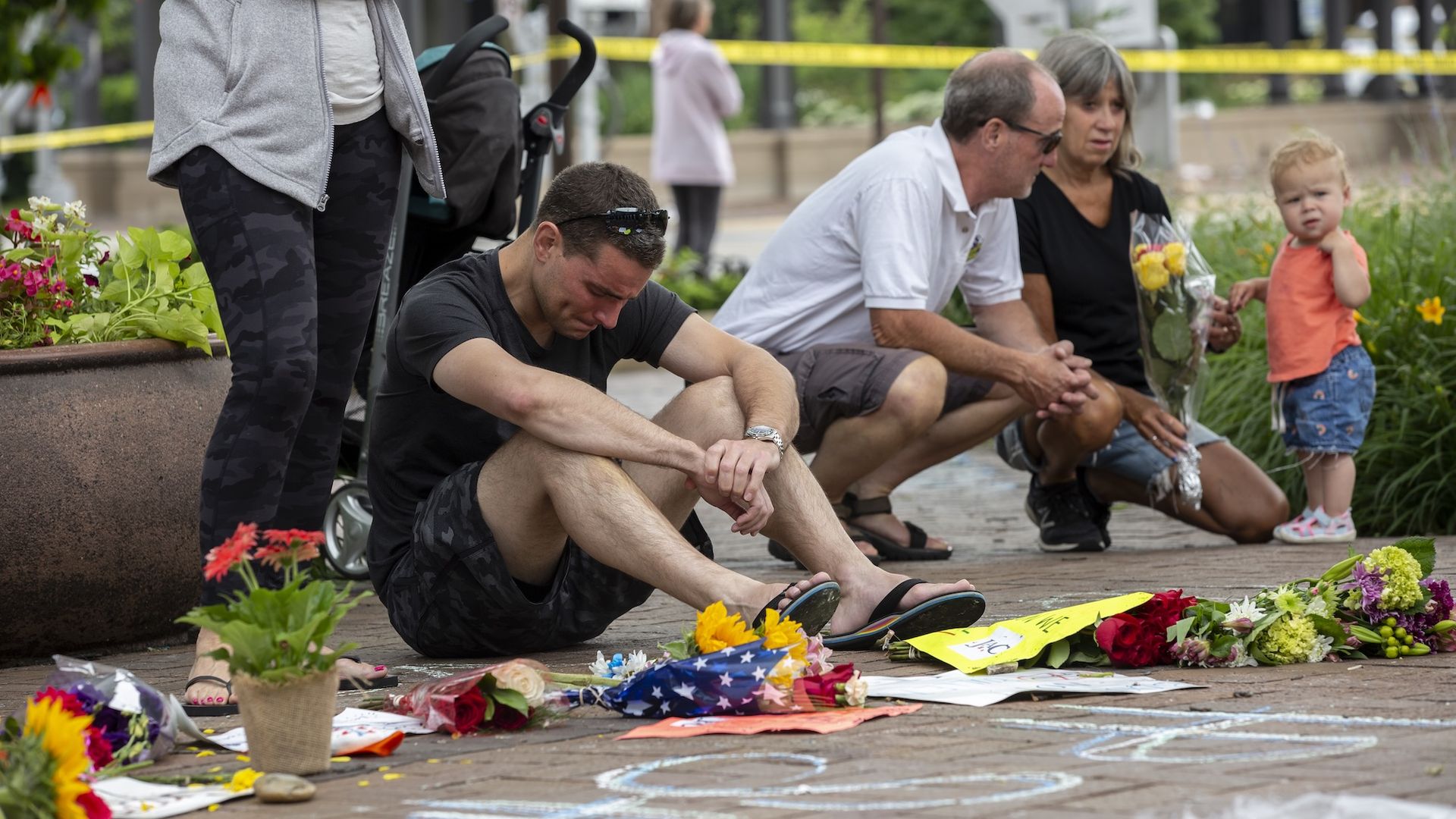 A man sits on the sidewalk with his head down in front of flowers with other people in the background.