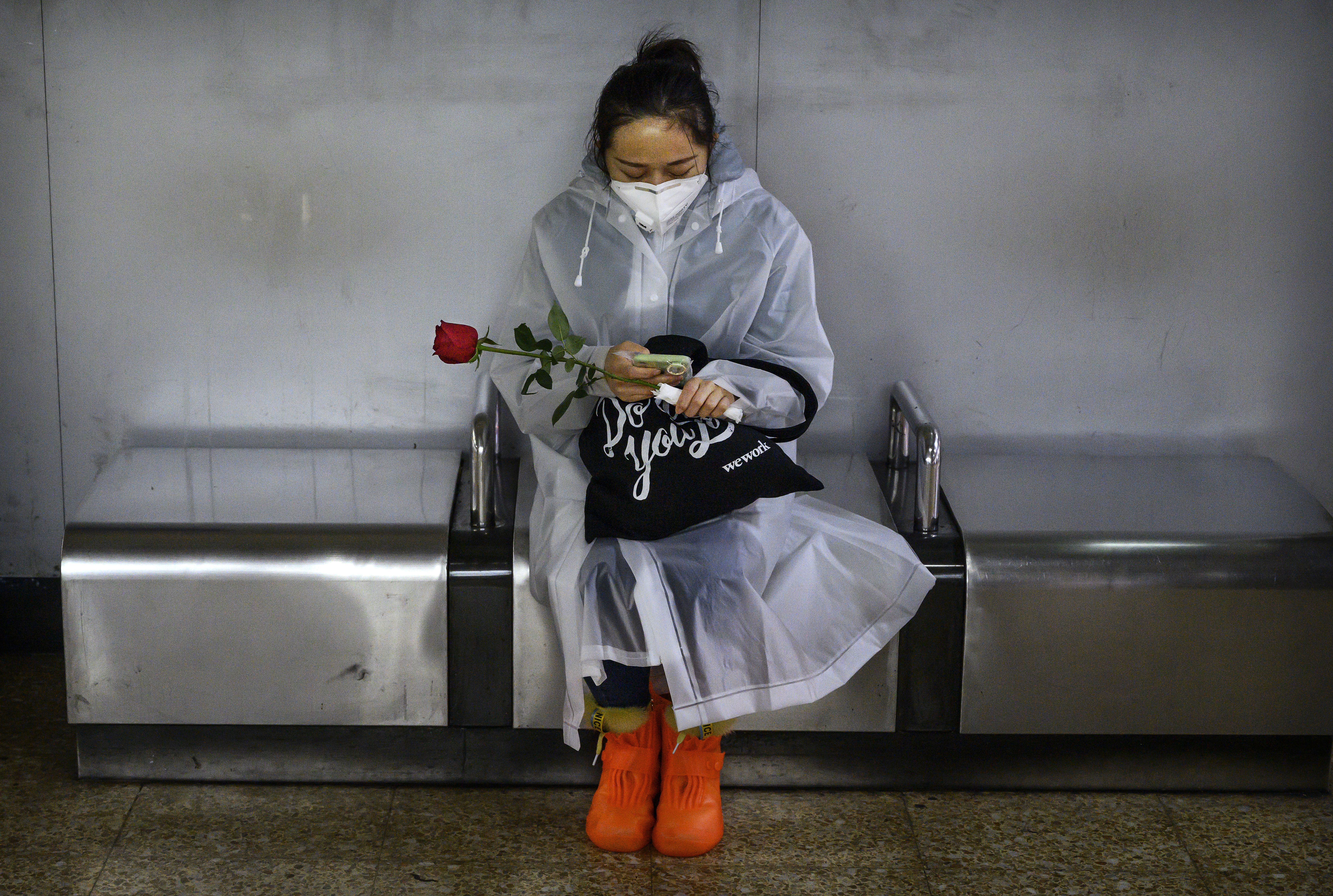 In this image, a woman in a protective face mask and poncho sits with a single red rose at a bus stop