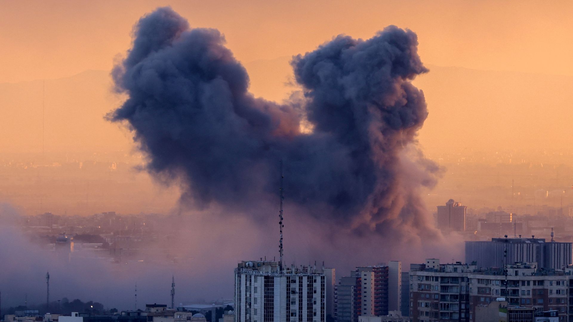 A plume of smoke rises from a city during sunset. The tones are blue and orange.