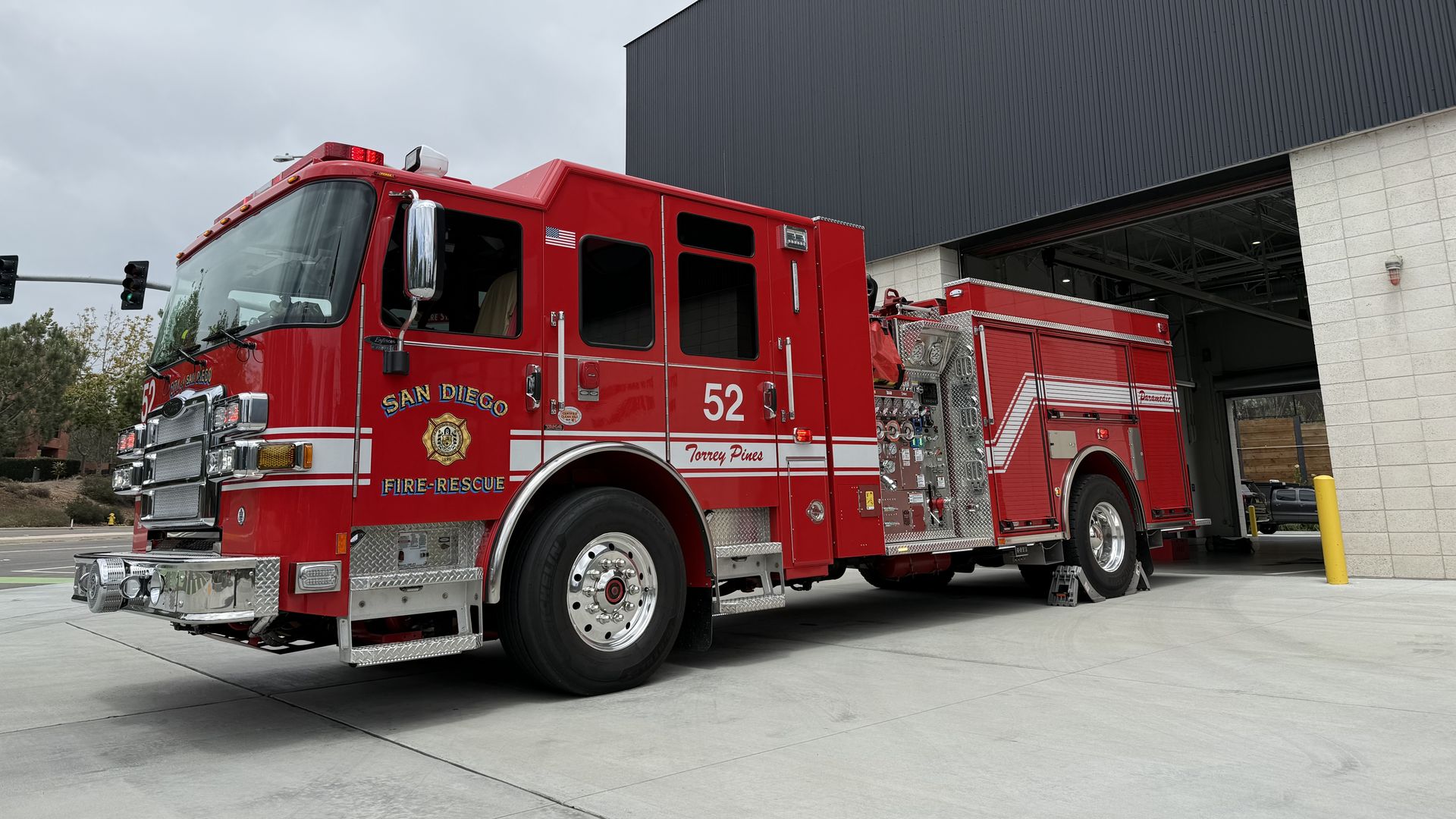 Red fire engine parked in front of a large dark gray industrial building under a cloudy sky; chrome details, ladders, and emergency lights visible, with San Diego Fire-Rescue branding.