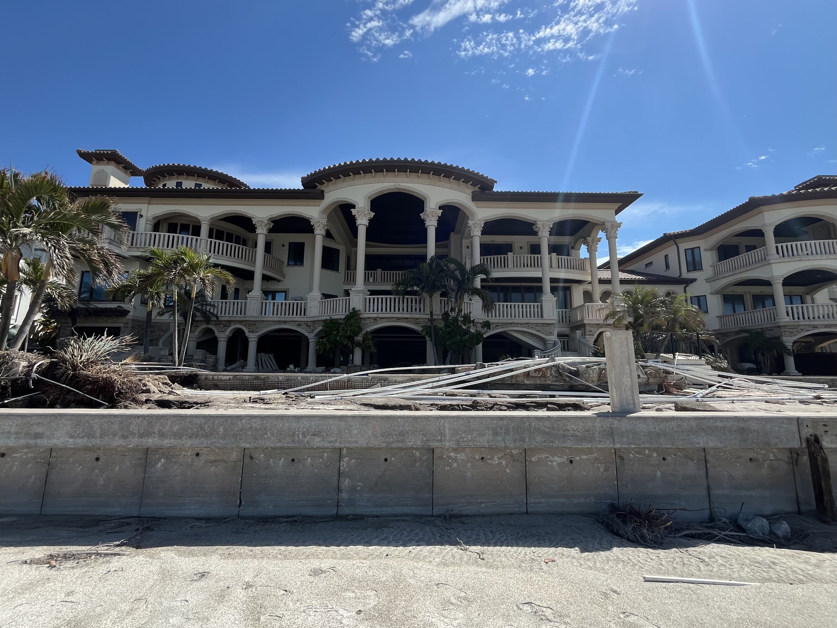 A tan stucco mansion with ornate columns and several balconies with a backyard littered with debris.