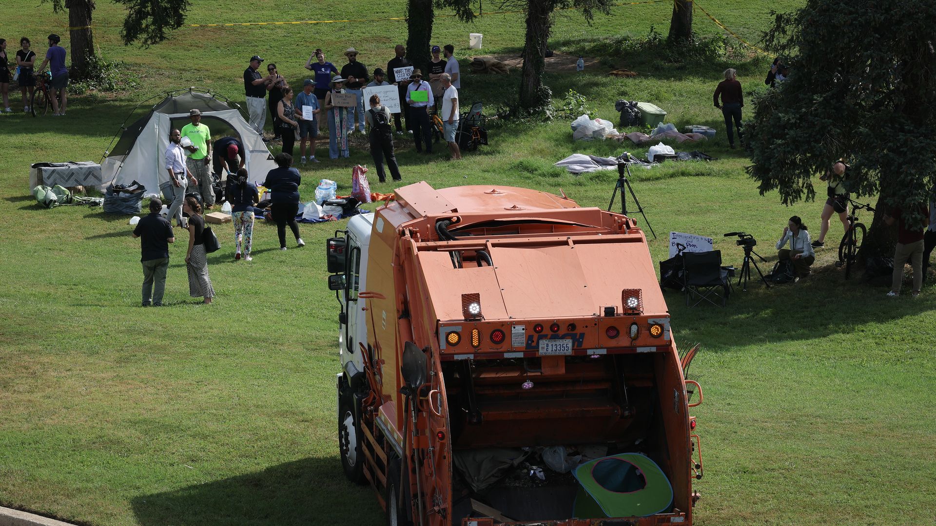 A garbage truck is parked on grass in front of a homeless encampment made up of tents. 
