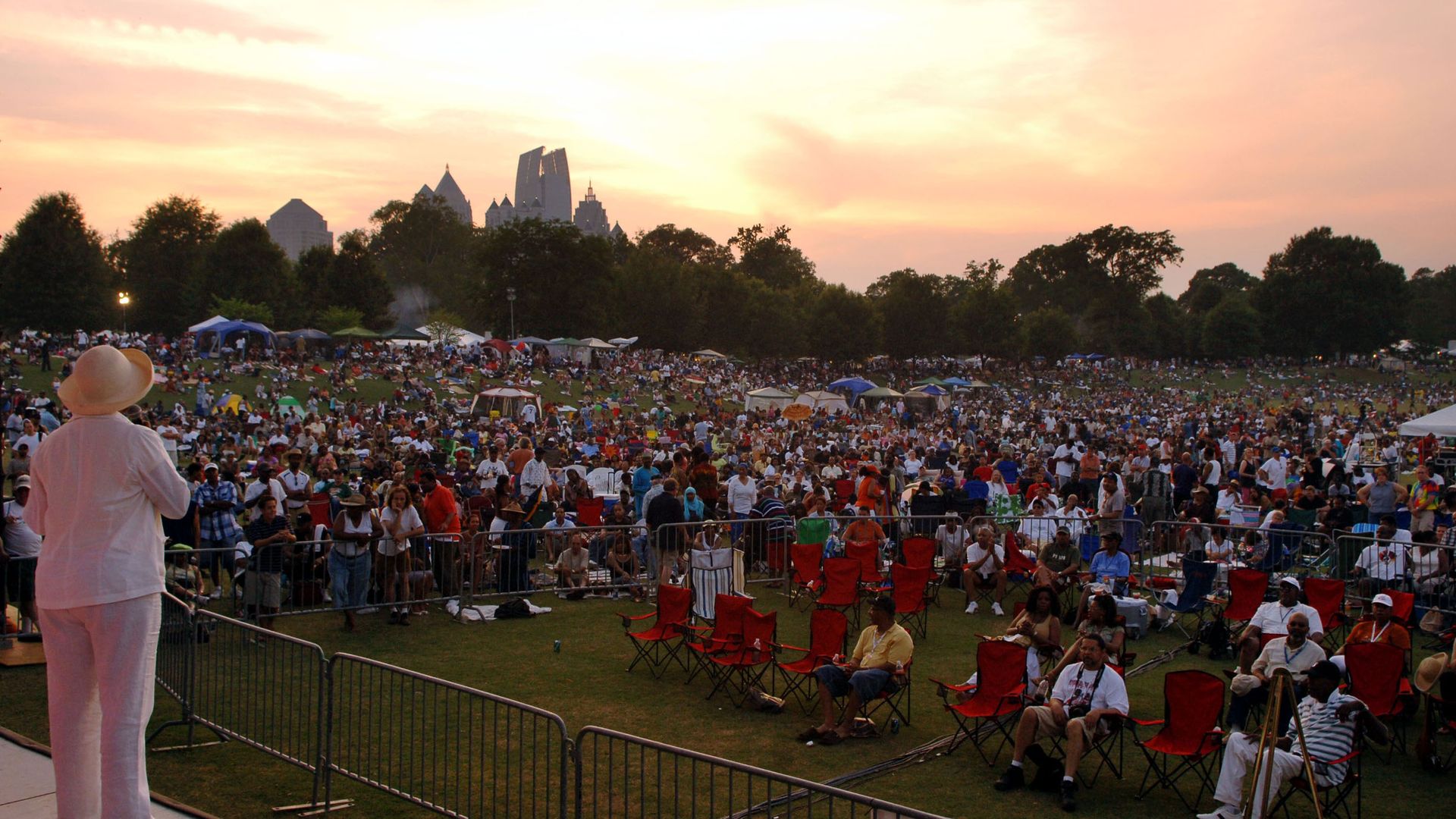 An individual wearing a hat addresses a massive outdoor crowd in a park as the sun sets behind the Midtown Atlanta skyline