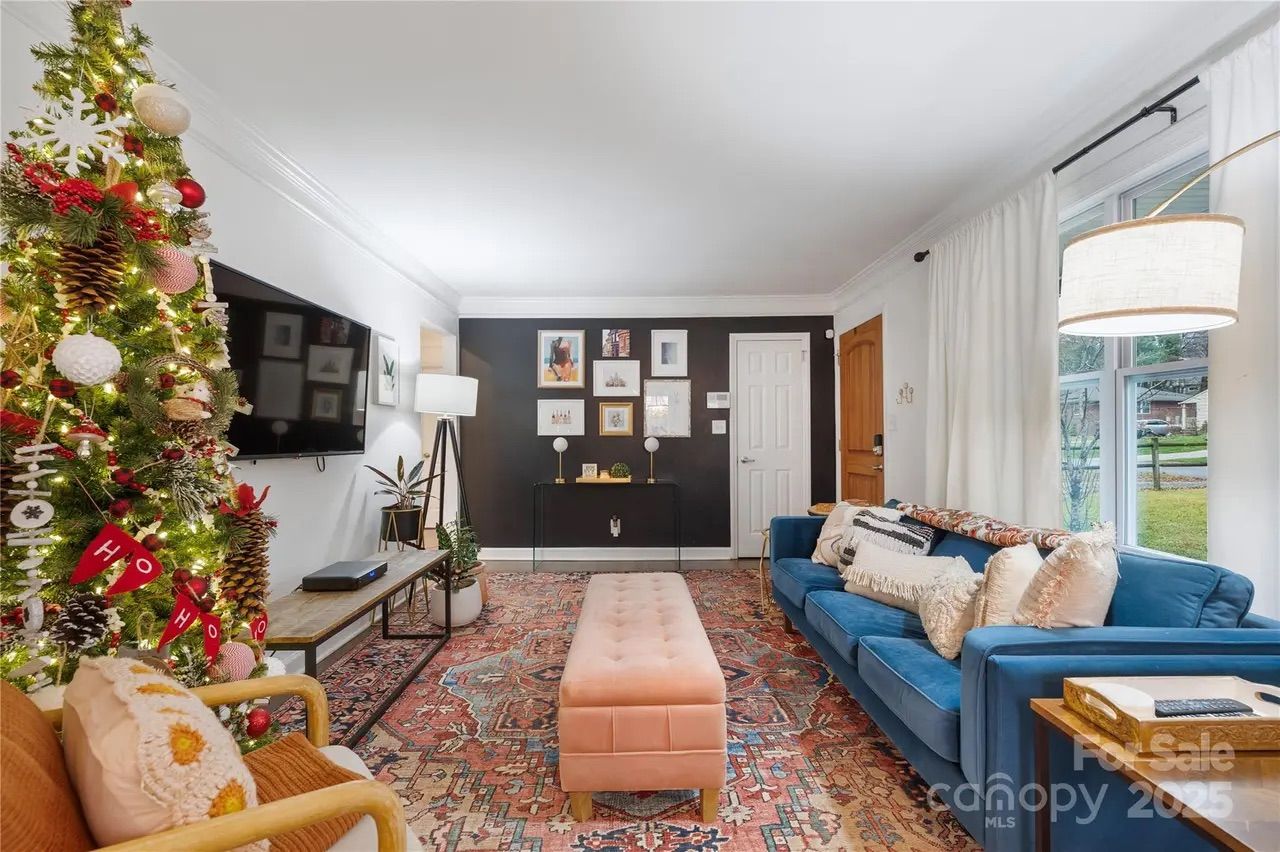 Cozy living room with a decorated Christmas tree, blue sofa with cushions, pink tufted ottoman, patterned rug, wall-mounted TV, black accent wall with framed art, and large window with white curtains.