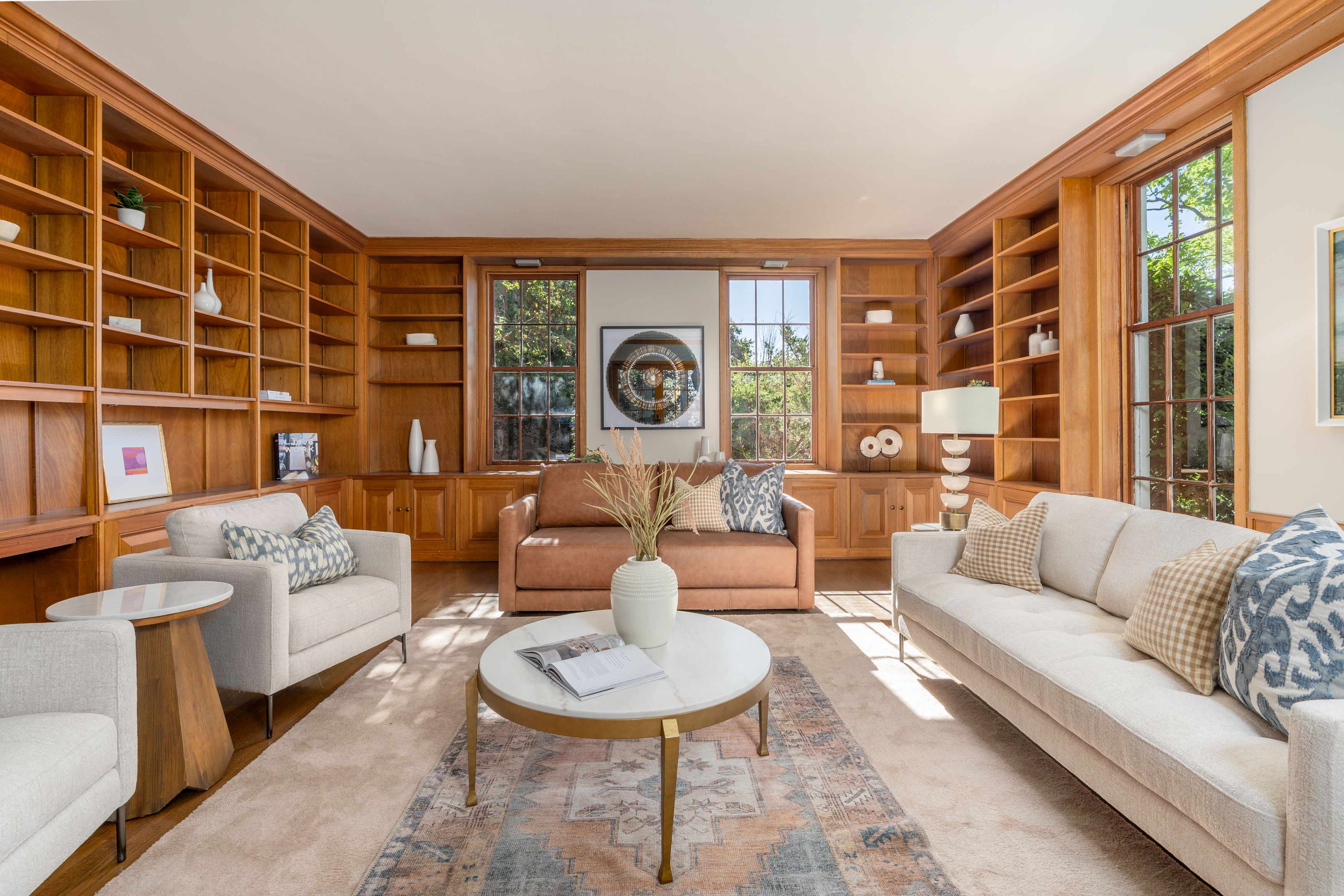 Bright living room with floor-to-ceiling wooden built-in shelves on two walls, beige carpet, and cream fabric sofas around a tan leather sofa. Round white coffee tables, large windows, and artwork.