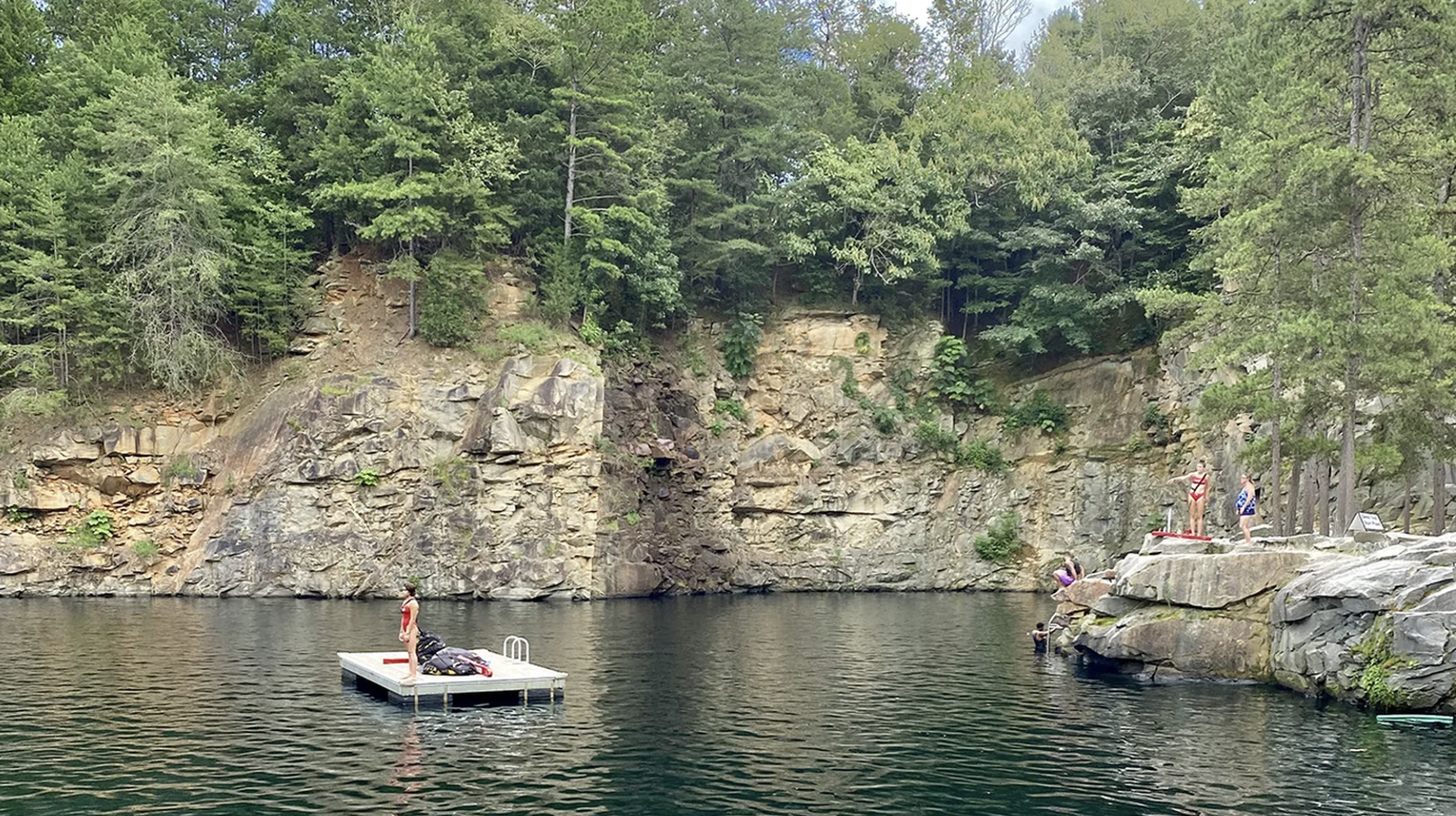 A natural swimming area with a rocky cliff surrounded by green trees. A person in a red swimsuit stands on a floating dock, while others prepare to jump or swim from rocks.