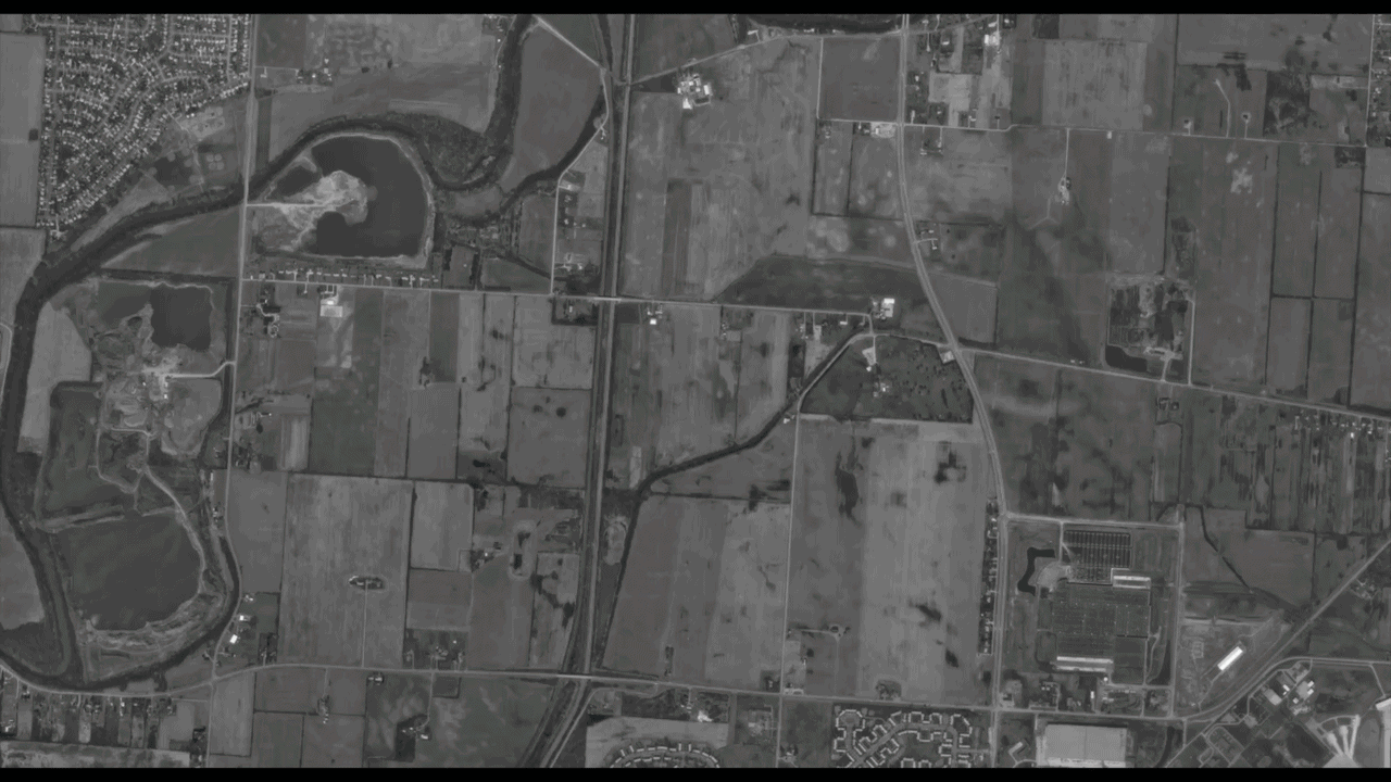 Grayscale aerial view of a rural area: a winding river with bends, patchwork fields, scattered houses, and a network of roads; a few farm or industrial buildings appear in the bottom-right.